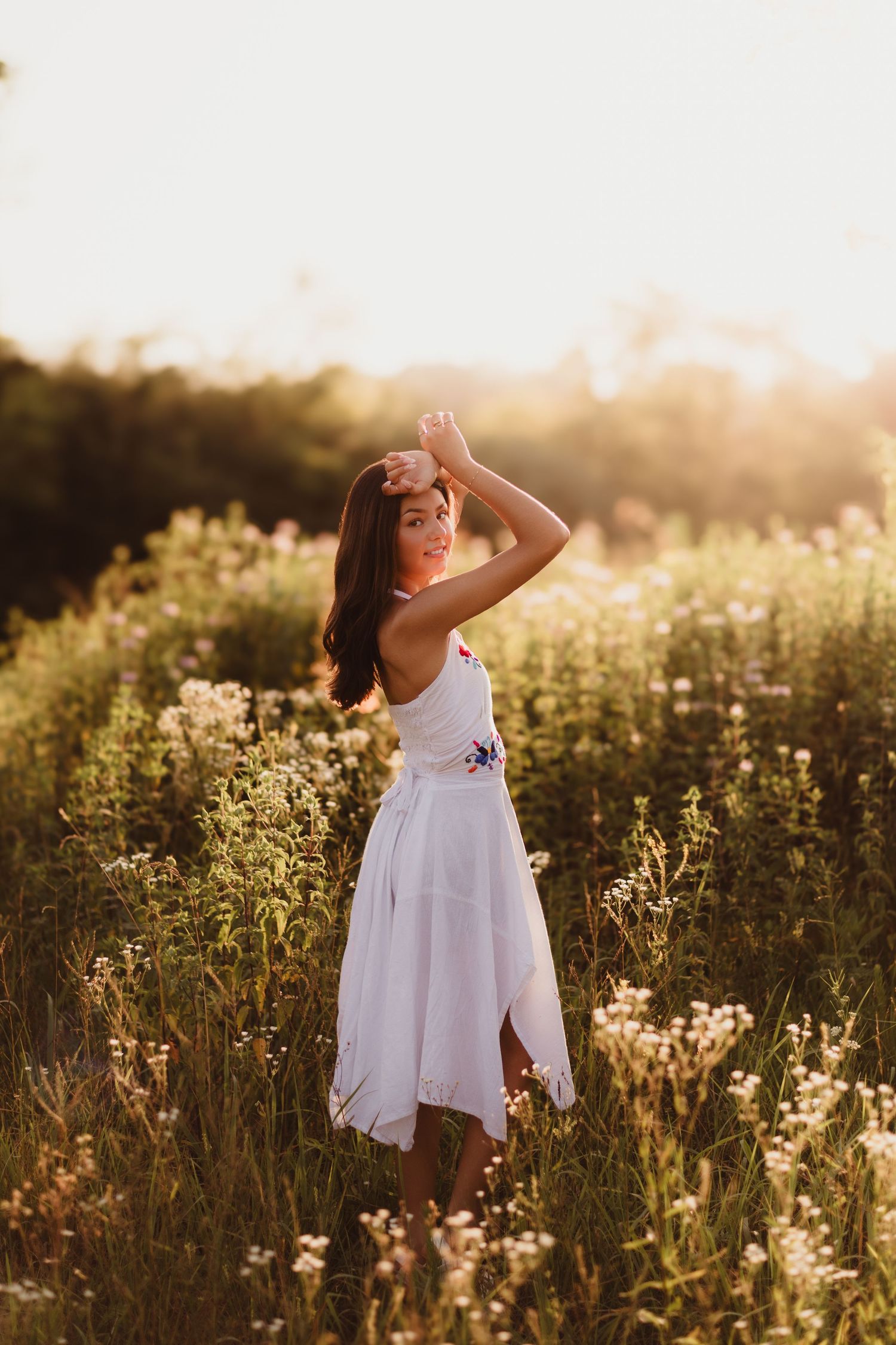 Young woman in white floral dress standing in golden hour sunlight among wildflowers in a meadow.