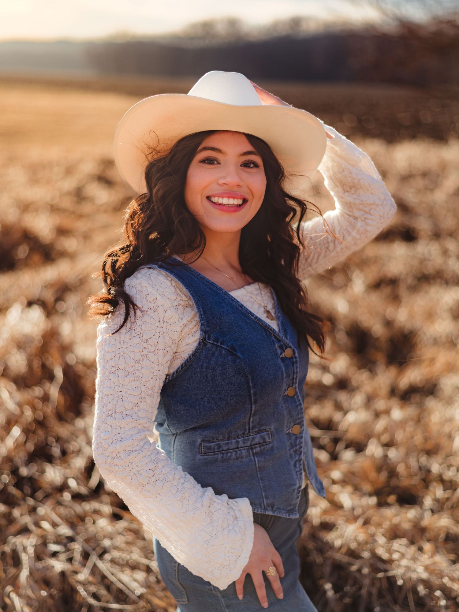 A person wearing a denim vest and white sweater poses playfully in a sunlit field during golden hour.