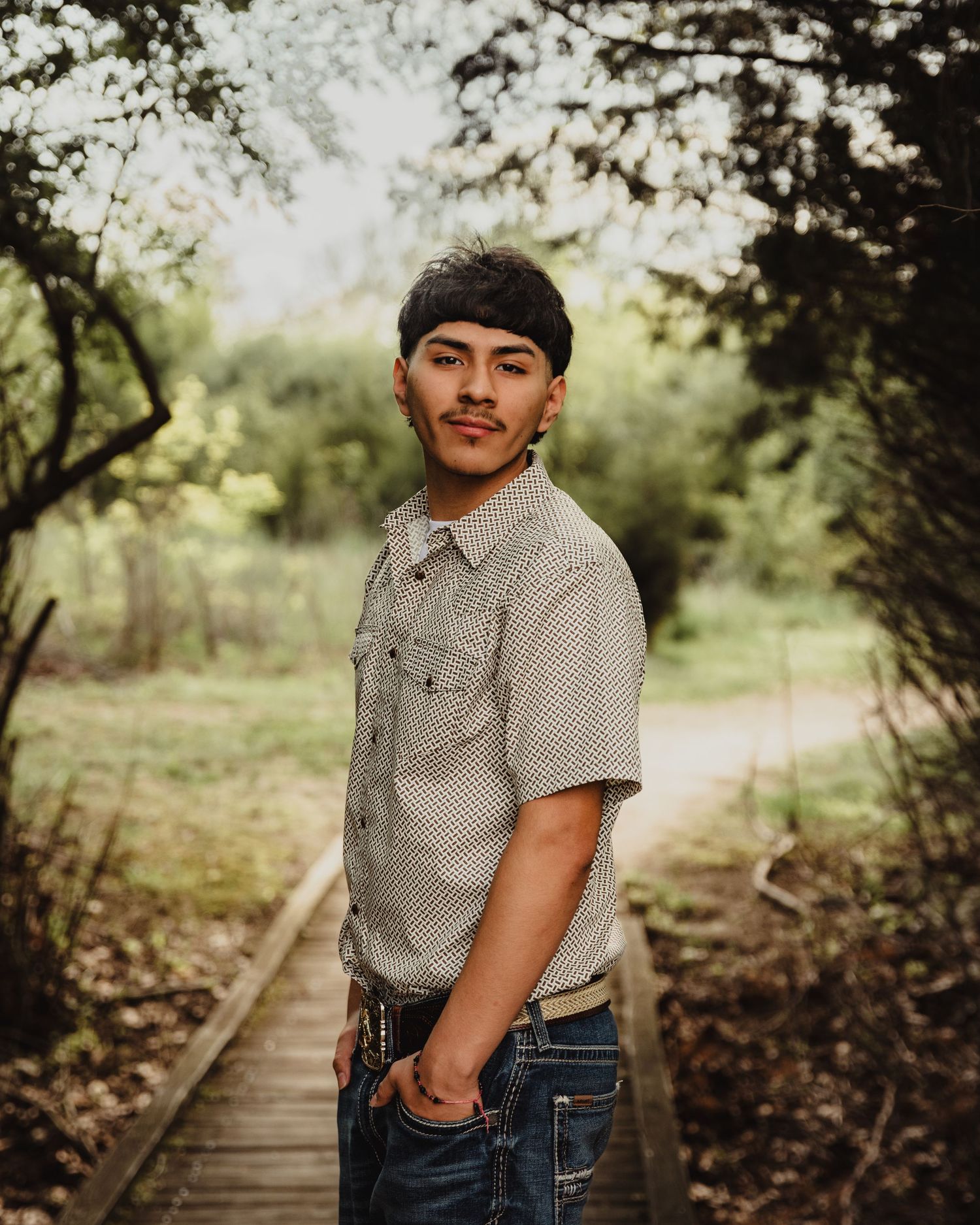 Young man in a patterned shirt and jeans poses on a wooden bridge path surrounded by lush green trees.
