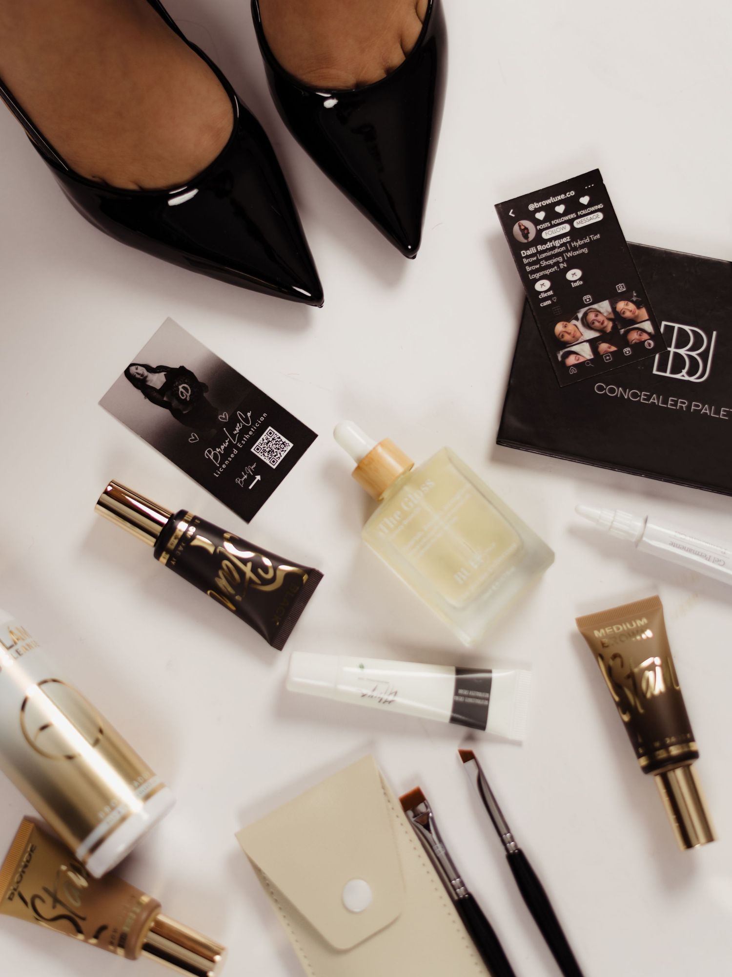 Flatlay of luxury makeup products, brushes, concealer palette, face oil, and black patent heels on white background.