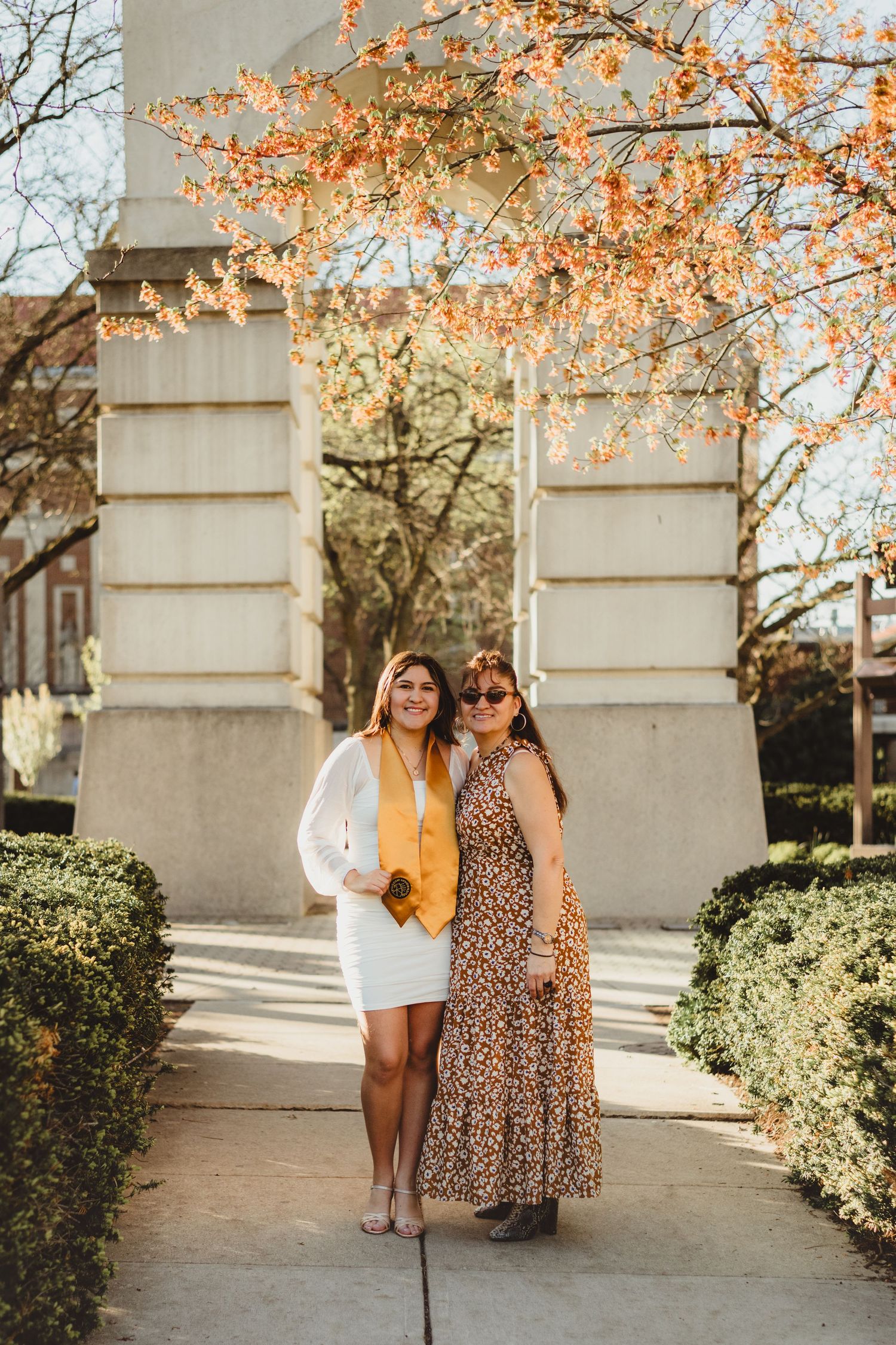 Two friends pose together in front of stone pillars on a sunny autumn day with orange foliage overhead  at Purdue Campus in West Lafayette.