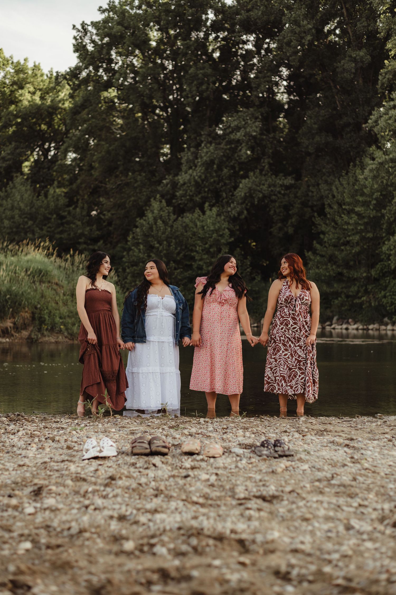 Four young women in elegant dresses stand barefoot holding hands near a peaceful riverside with tall trees.
