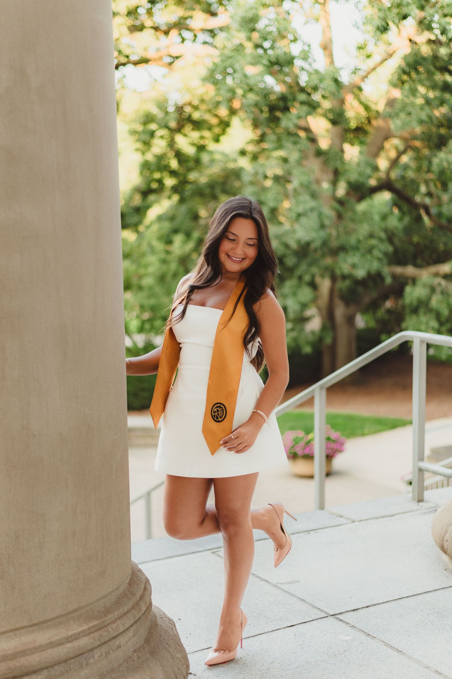 A person in a white dress and brown vest poses playfully between stone columns on a brick walkway  at Purdue Campus in West Lafayette, IN