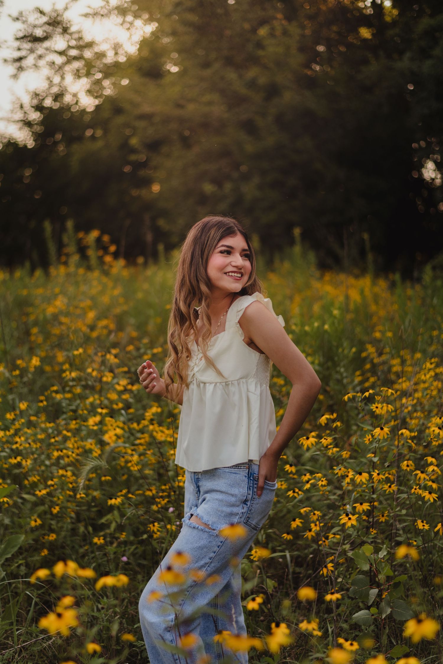 Person in white ruffle top and denim jeans laughs joyfully while standing in a field of yellow wildflowers at sunset.
