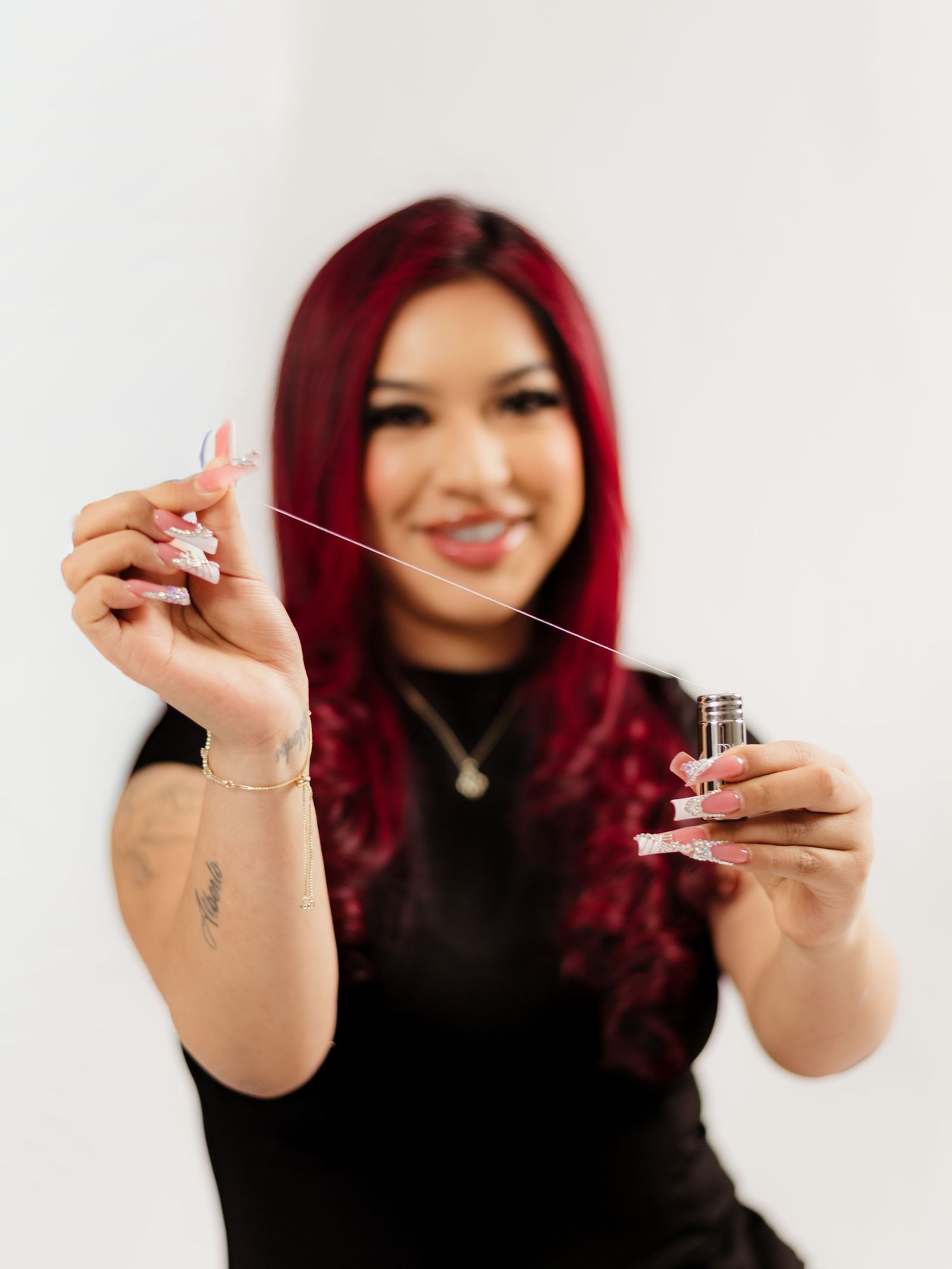 Woman with red hair holding dental floss and a small bottle, smiling, wearing a black top with tattoos visible.