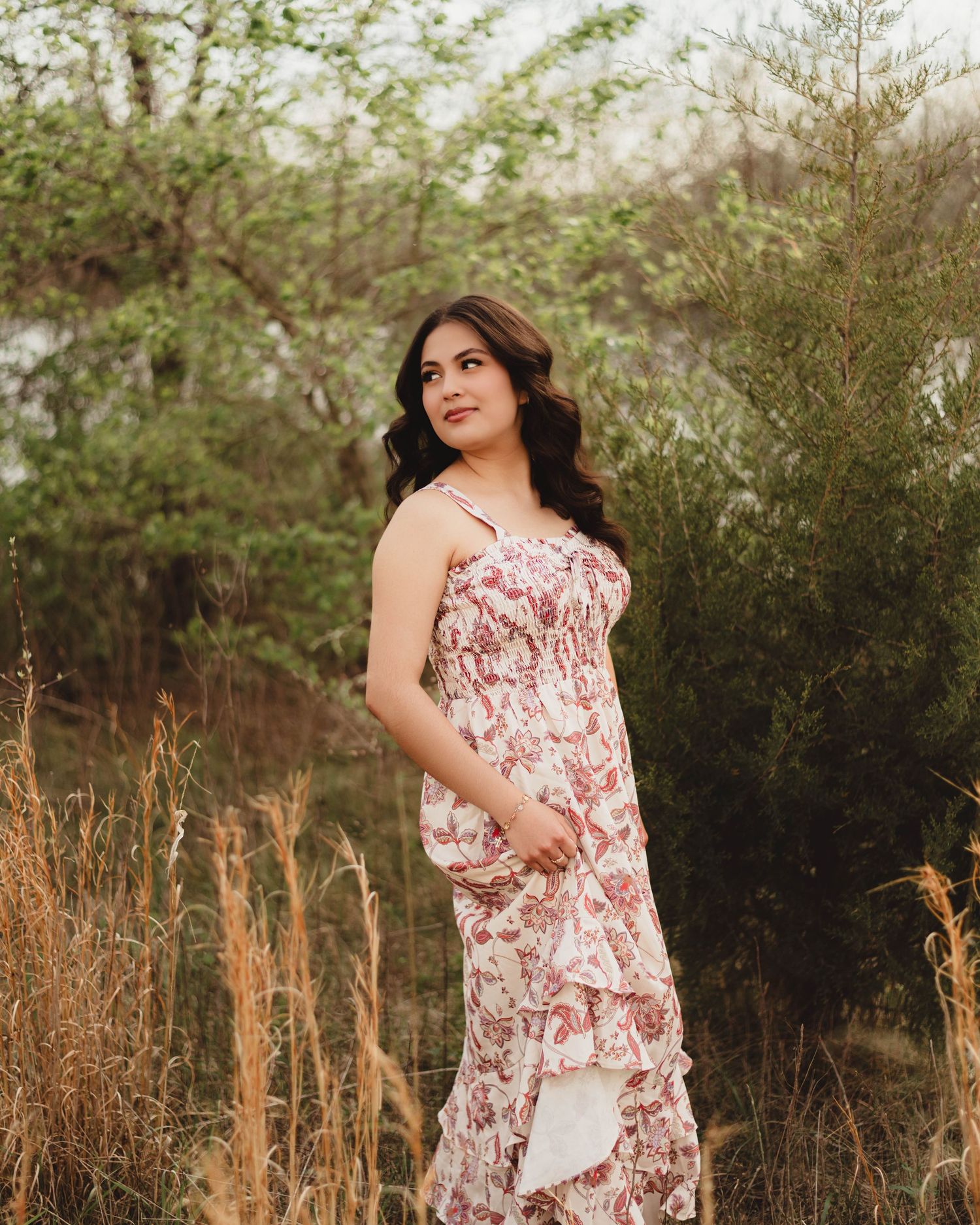 A young woman in a floral ruffled maxi dress poses gracefully outdoors among tall grass and lush green trees in Lafayette, indiana