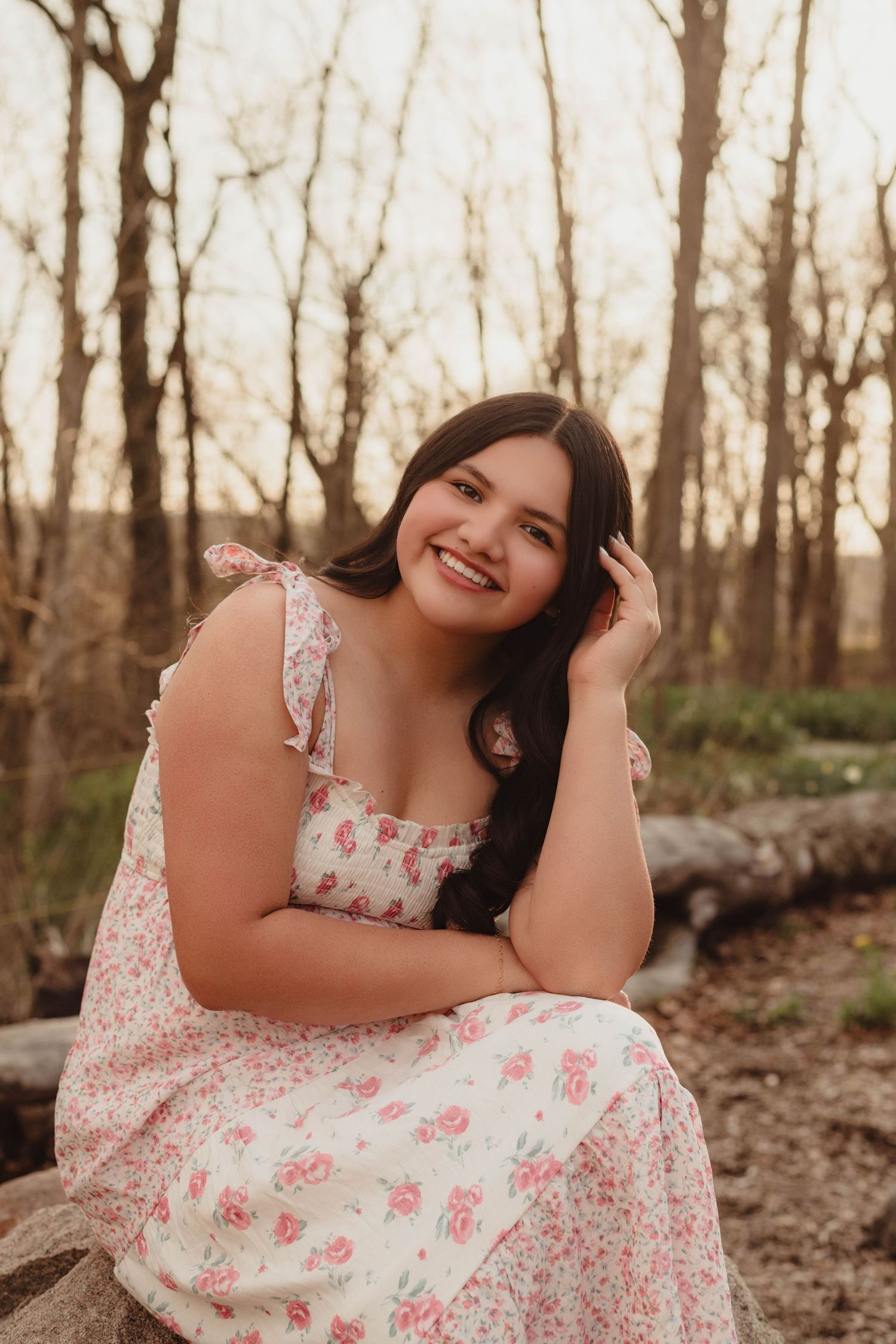 High school senior in floral dress sitting outdoors, smiling warmly among bare trees in golden hour light in Lafayette, Indiana.