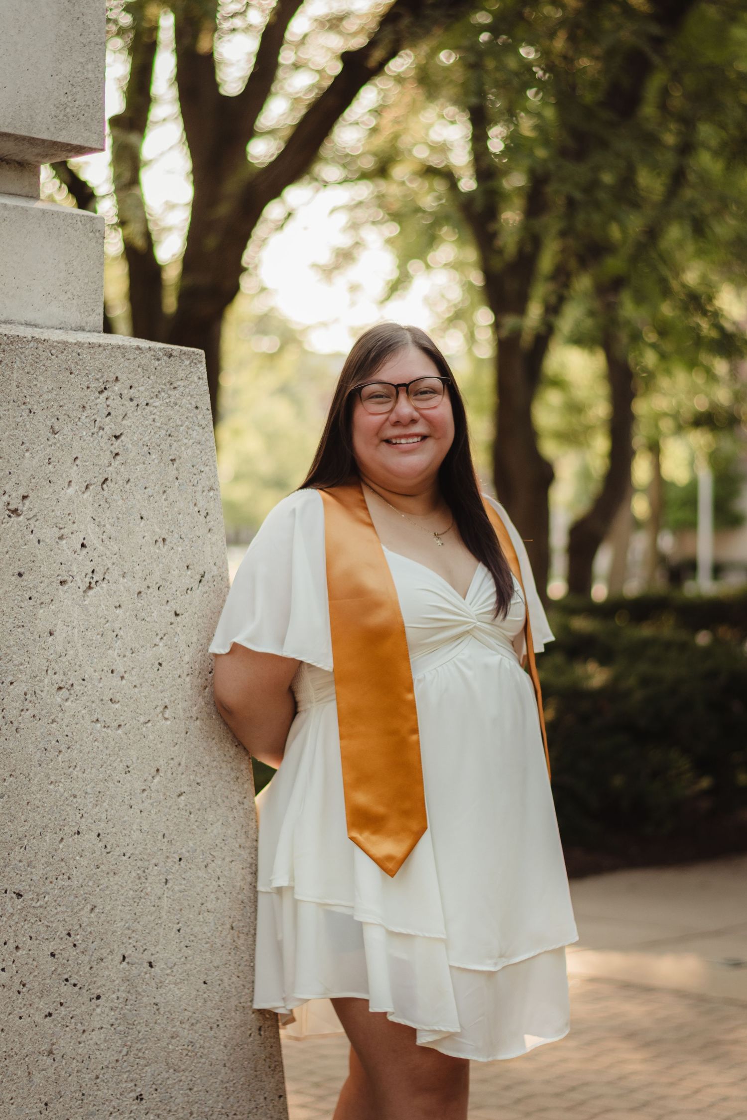 A graduate in a white dress and gold sash poses outdoors on campus with a bright smile.