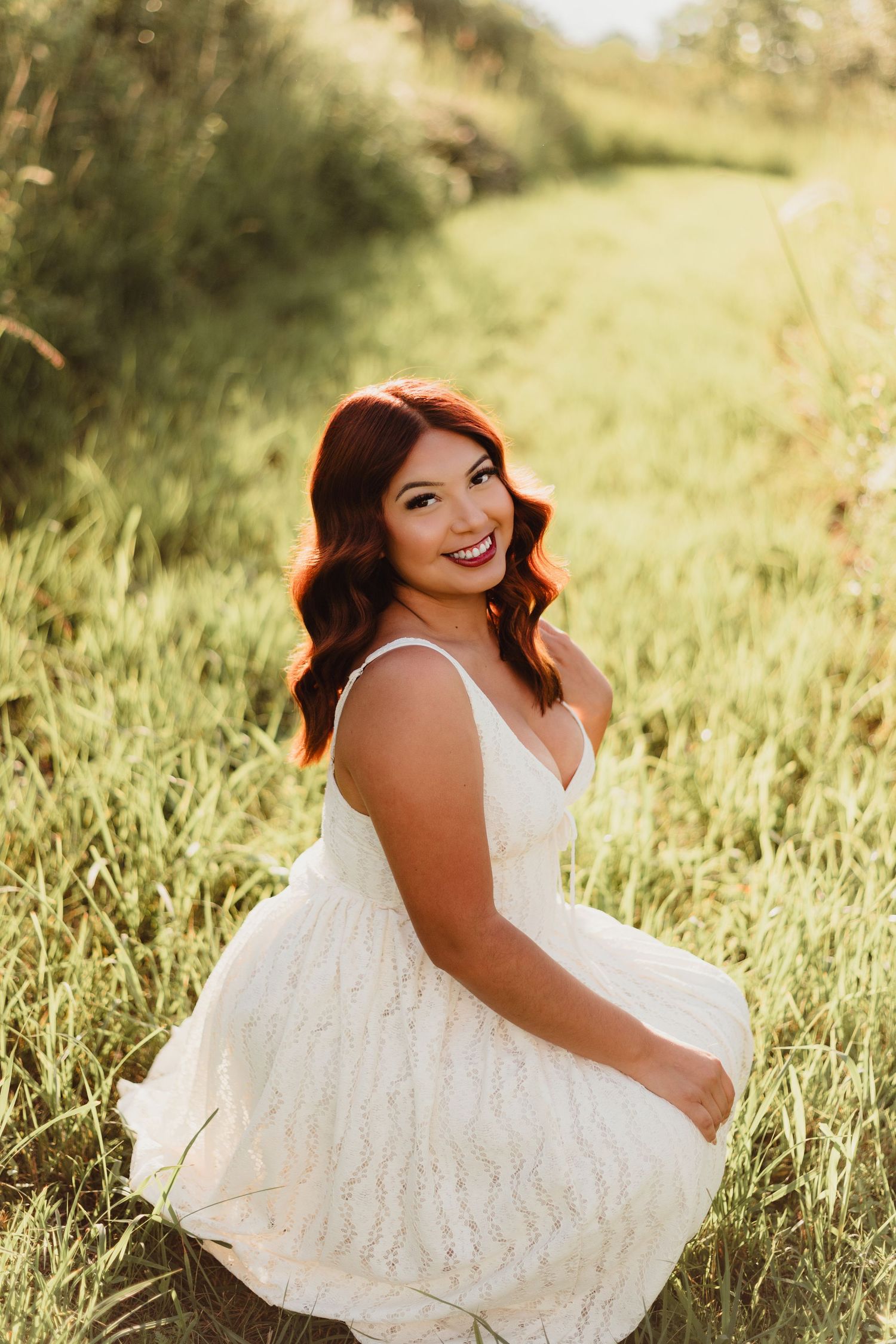 A young woman with auburn hair in a white lace dress smiles over her shoulder while sitting in a sunlit green meadow.