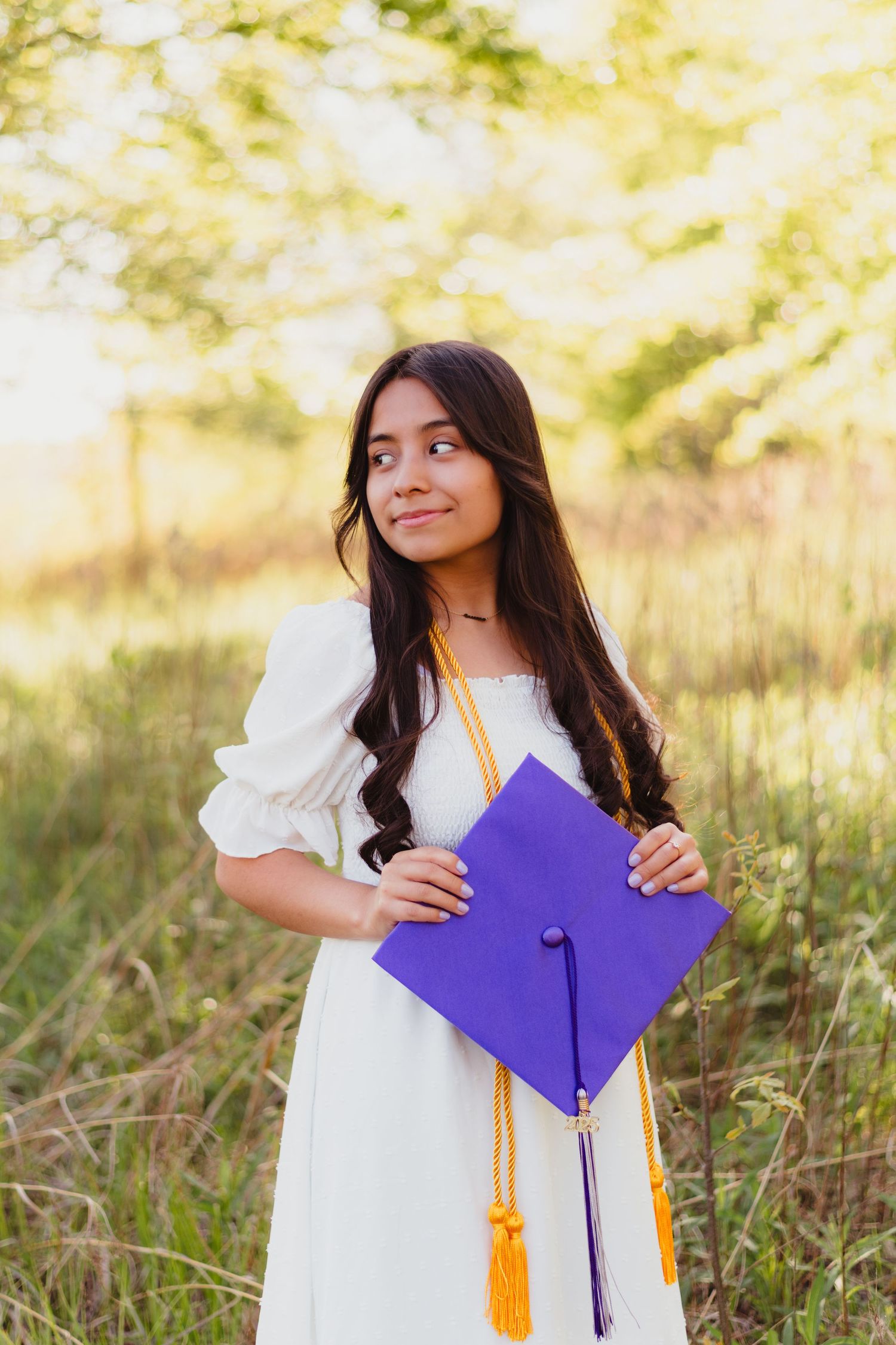 Graduate in white dress holds purple graduation cap with gold tassel while posing outdoors in sunlit field.