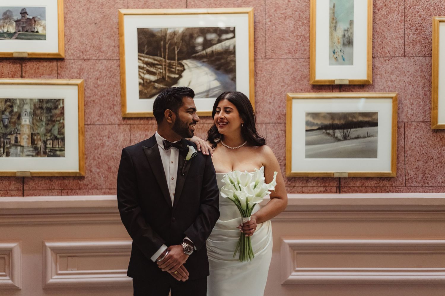 A romantic wedding couple shares an intimate moment in an elegant gallery space with framed artwork on pink walls.