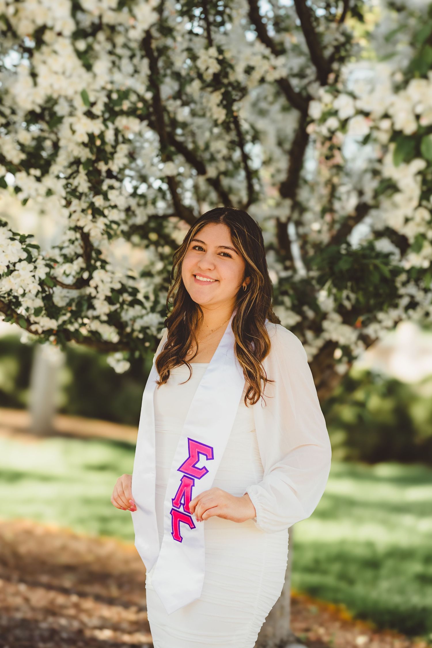 A person in a white sweater poses joyfully among blooming spring cherry blossom trees in a sunlit outdoor setting at Purdue Campus.