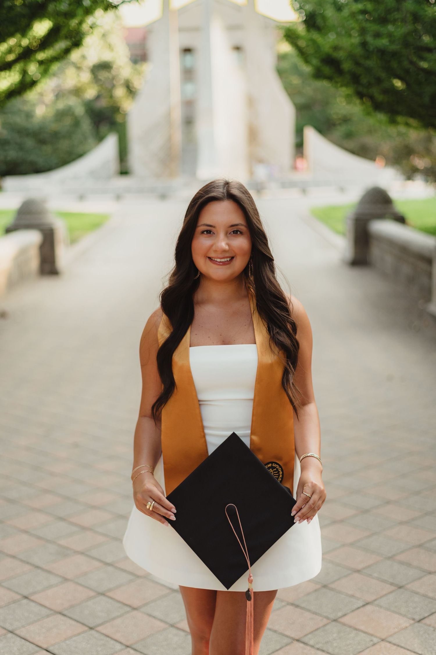 Graduate in white dress and brown sash holding graduation cap in outdoor campus setting  at Purdue Campus in West Lafayette, IN.