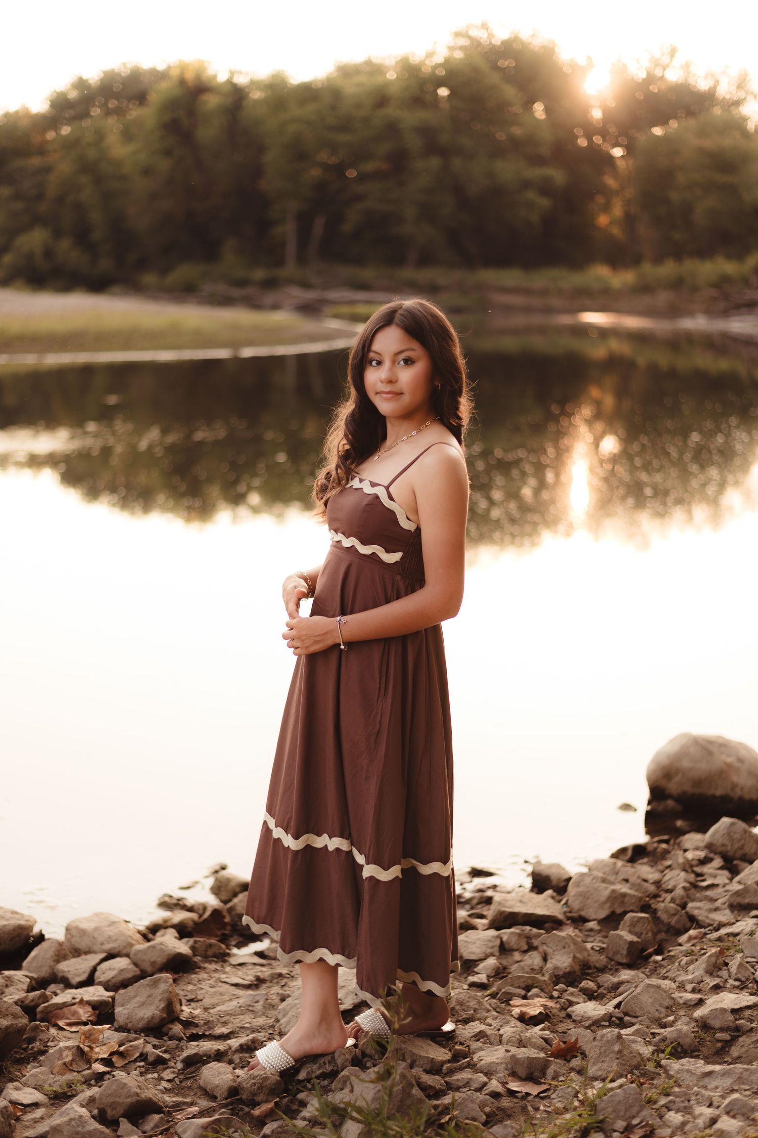 High School Senior in a brown dress sits on rocky shoreline at sunset in Lafayette, Indiana.