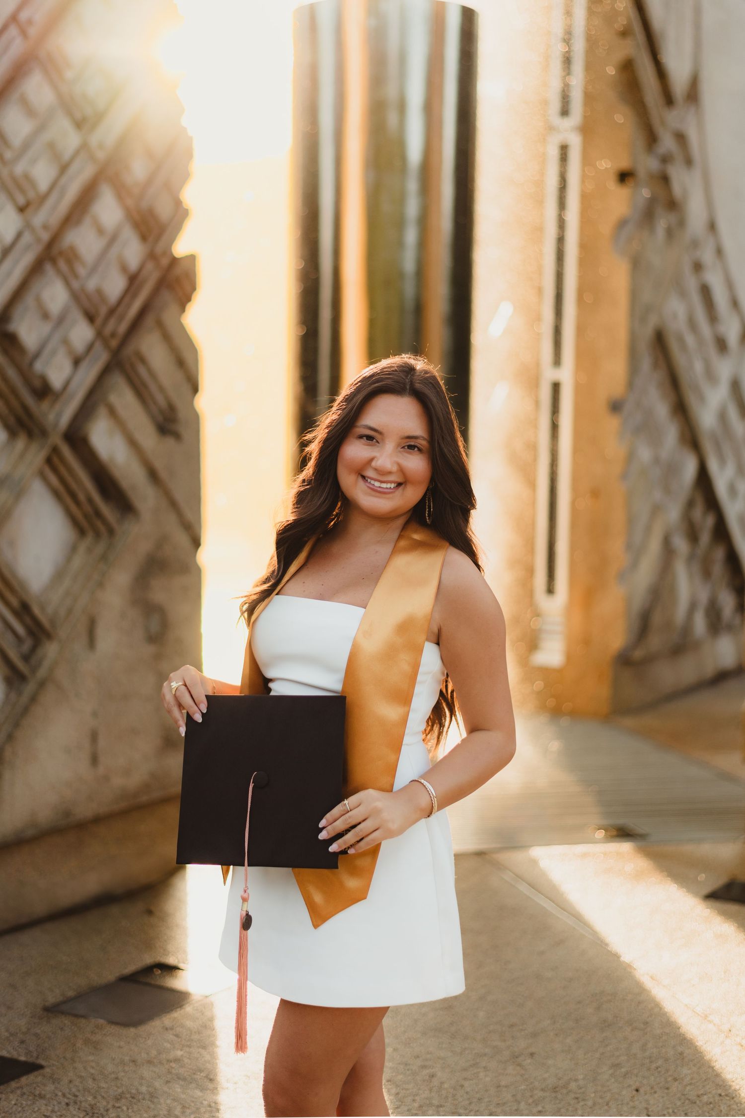 Graduation photoshoot in sunlit archway with white dress and brown academic stole at Purdue Campus in West Lafayette, IN.