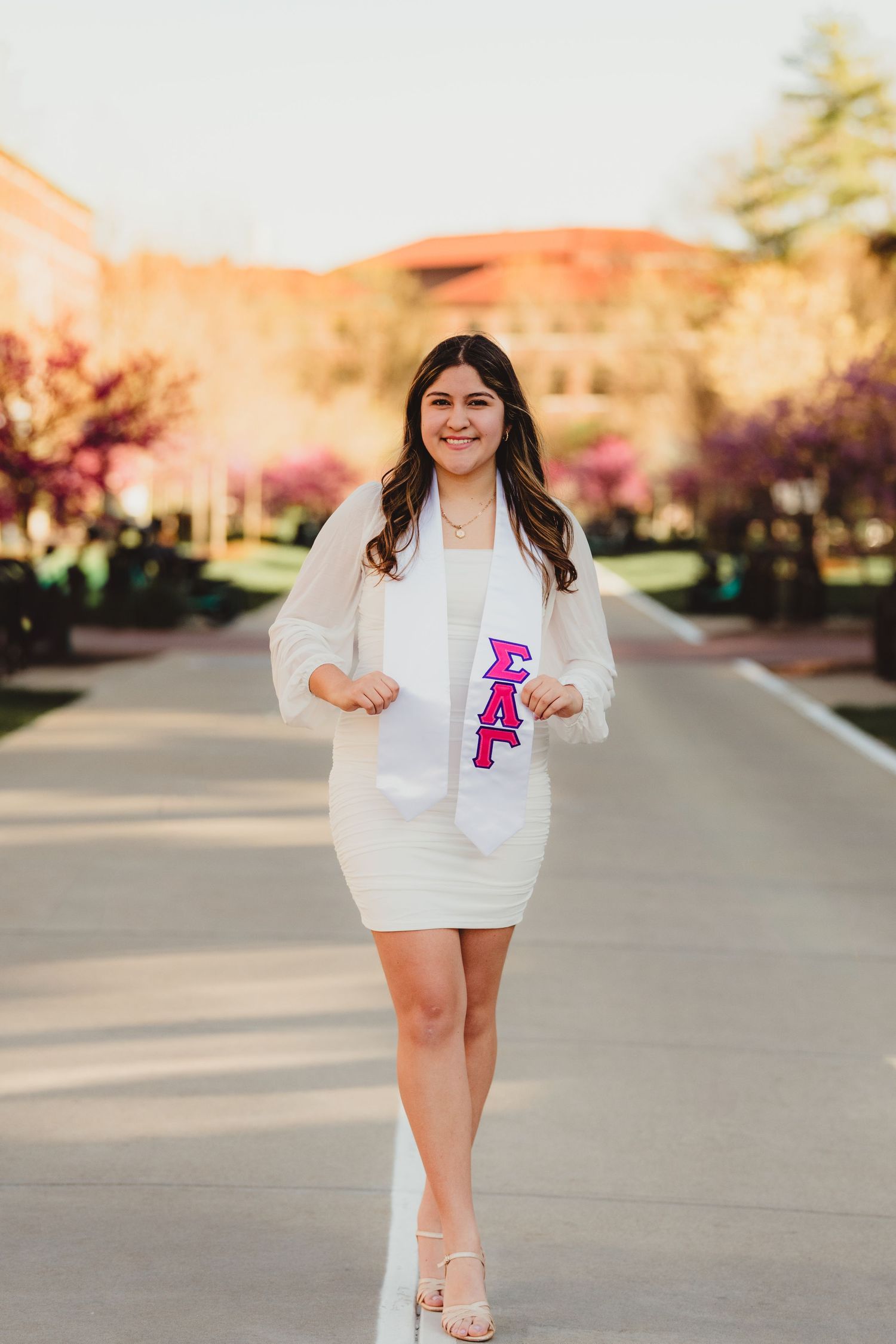 Purdue graduate poses in a white dress and blazer on a tree-lined pathway during golden hour at Purdue Campus in West Lafayette, IN