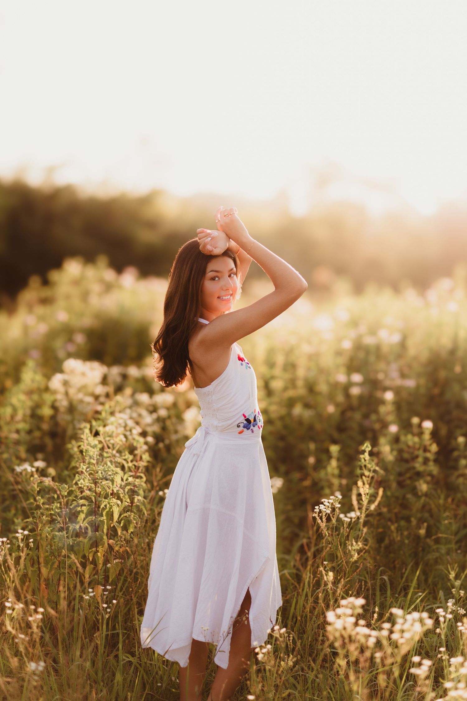 Young woman in white floral dress standing in a sunlit wildflower field, smiling over her shoulder at golden hour.