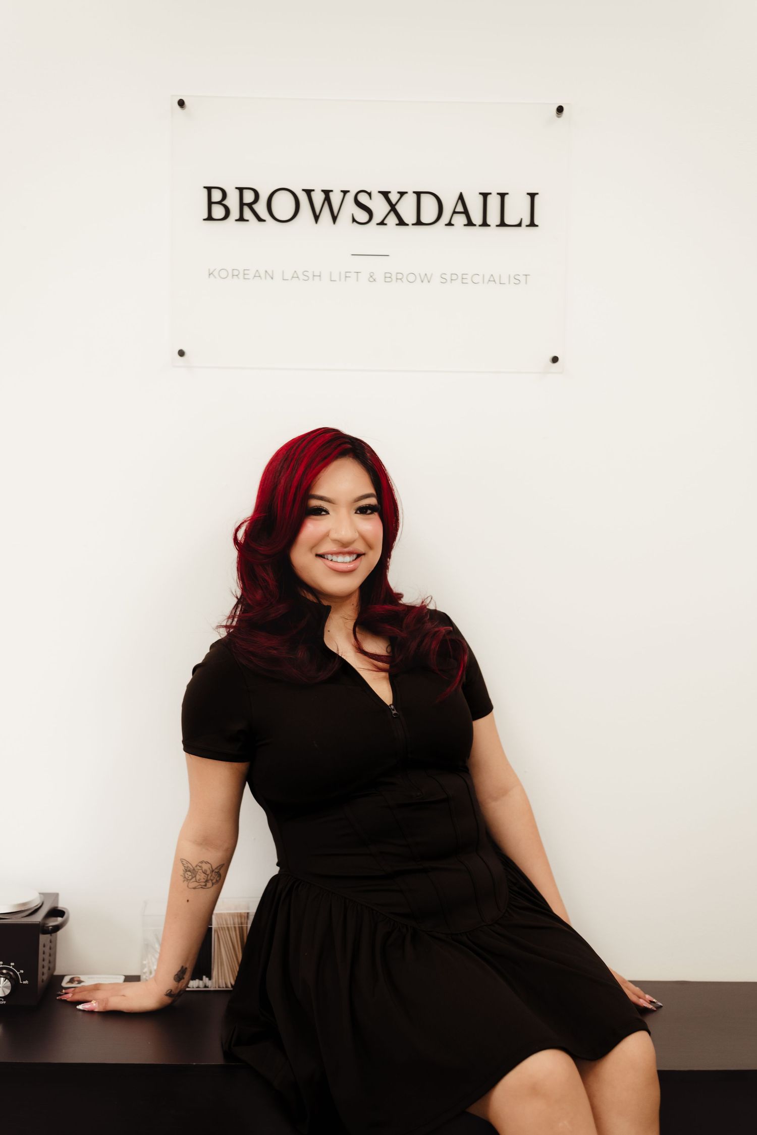 Woman with red hair in black dress sits before BROWSXDAILI Korean lash lift and brow specialist sign.