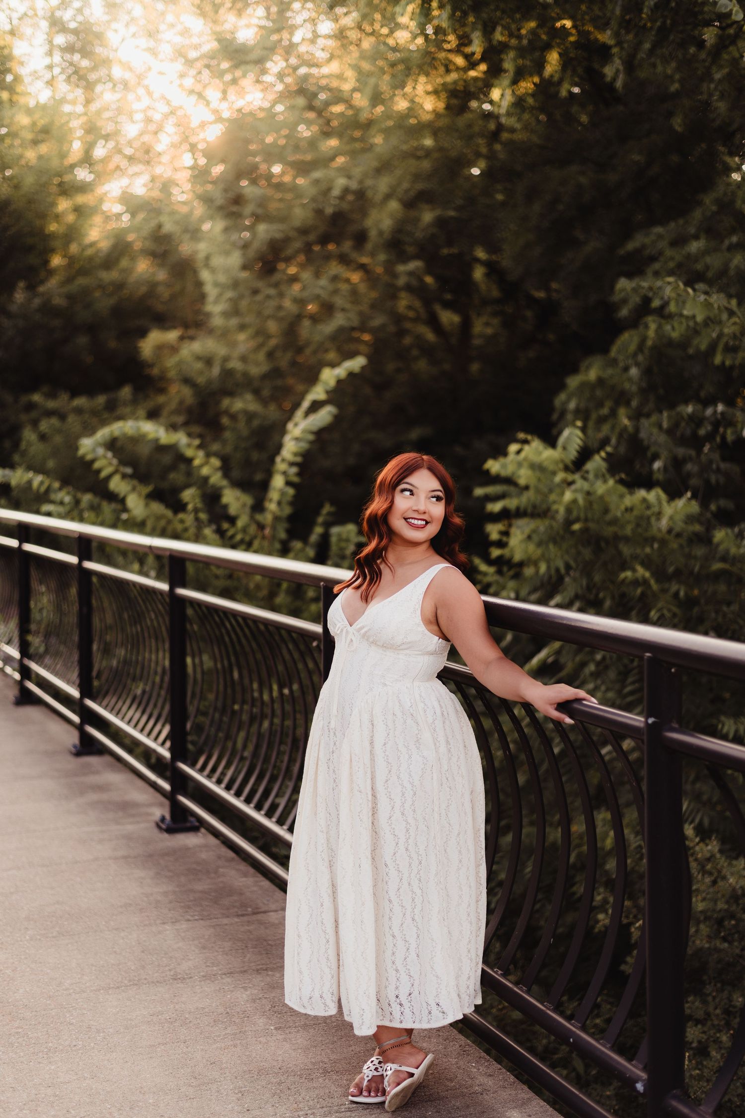 Woman in white lace midi dress posing on a bridge railing surrounded by golden sunlit greenery.