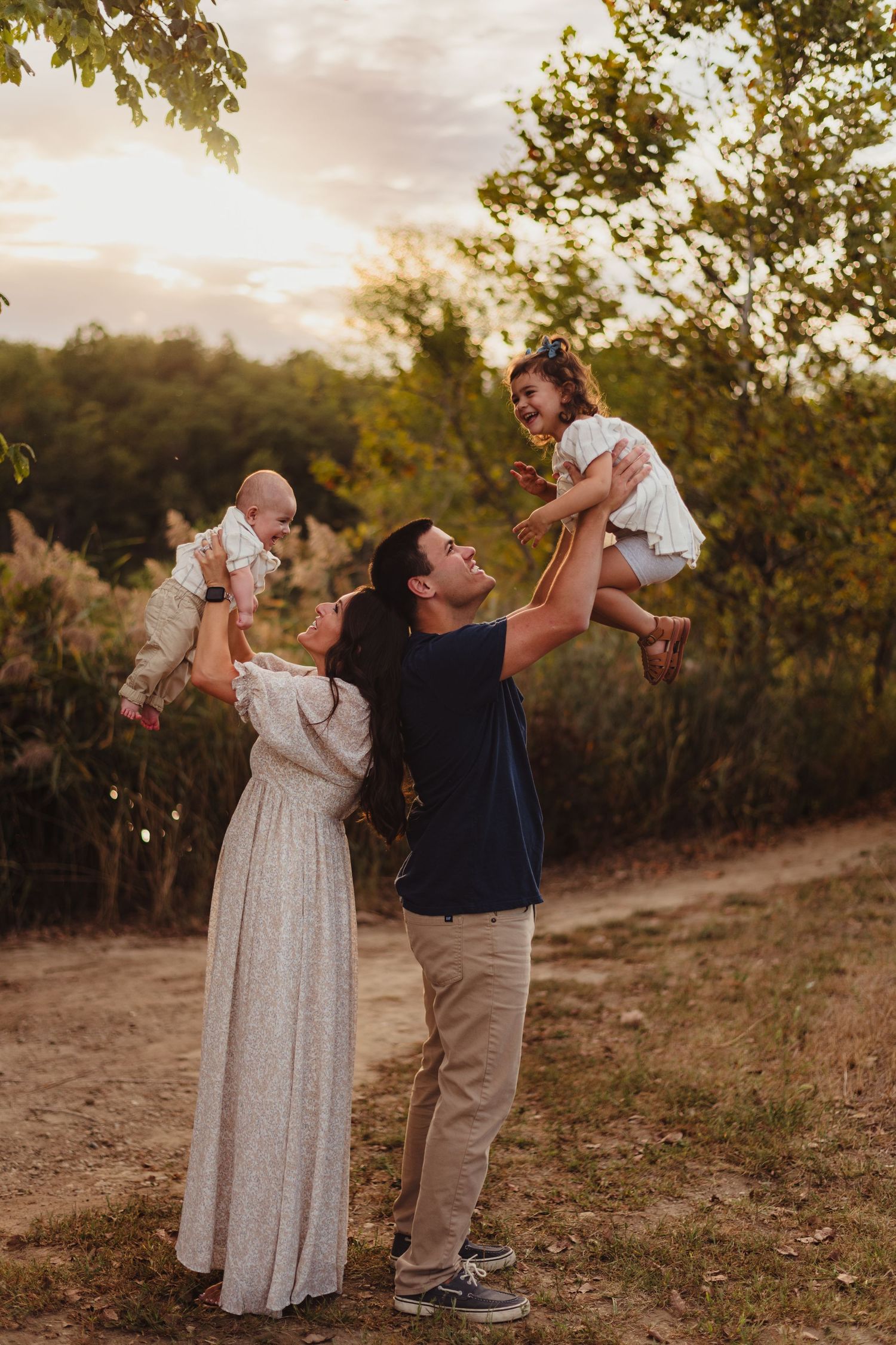 A joyful family plays outdoors at golden hour, parents lifting two young children in a lush green nature setting.