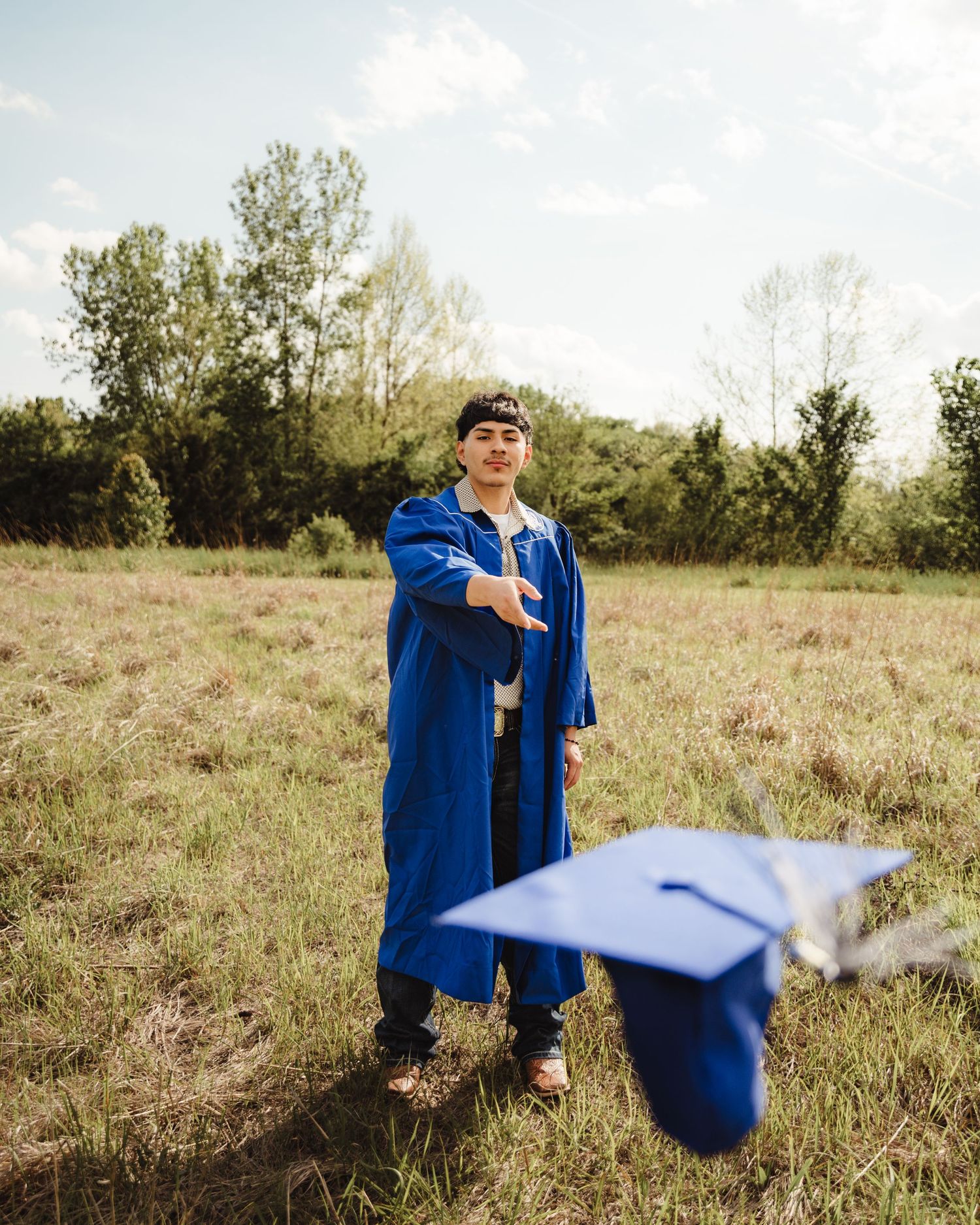 A graduate in blue cap and gown tosses their graduation cap in a green field outdoors.