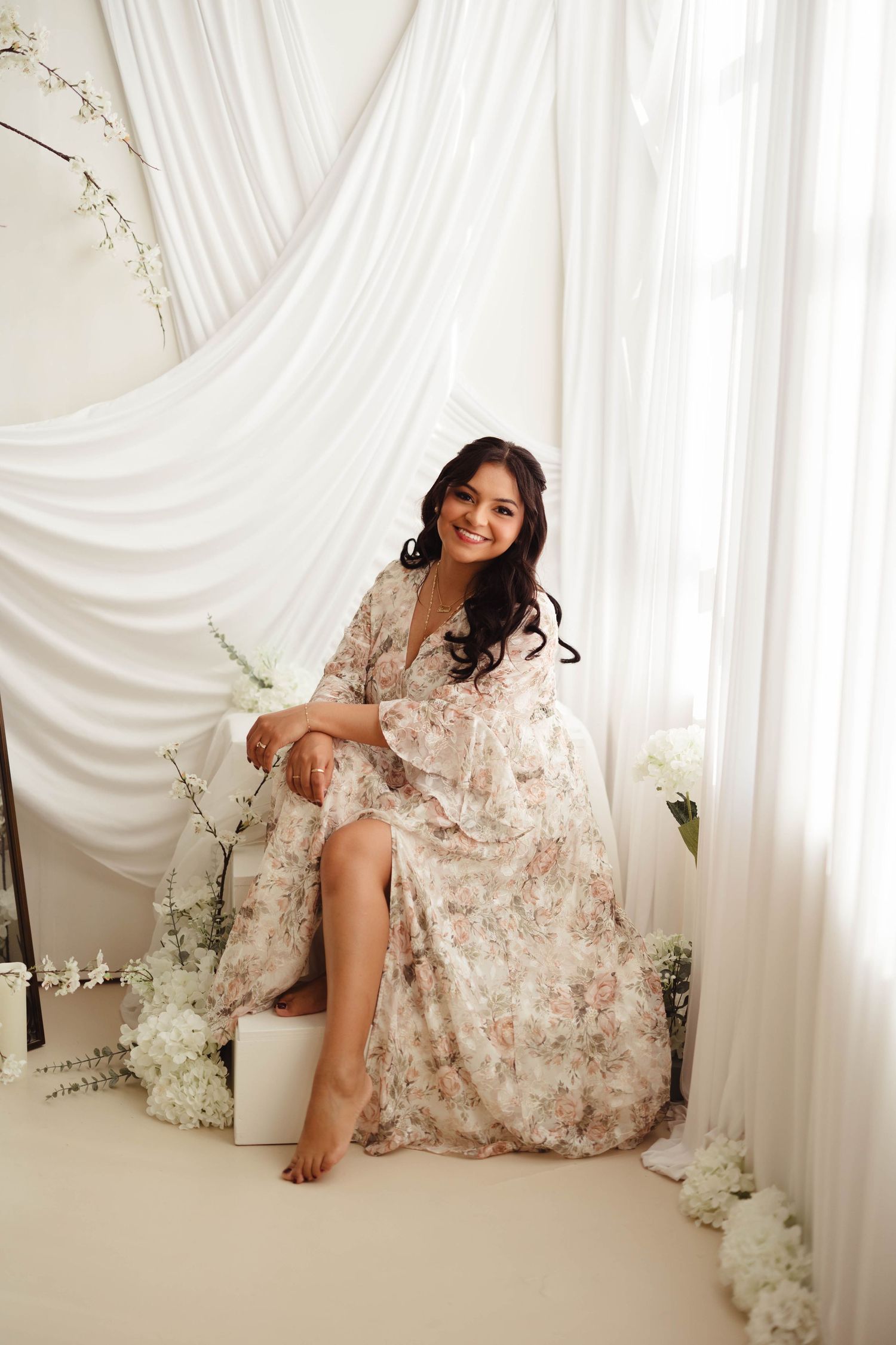 Smiling woman in floral maxi dress seated among white flowers and draped curtains in bright studio setting.