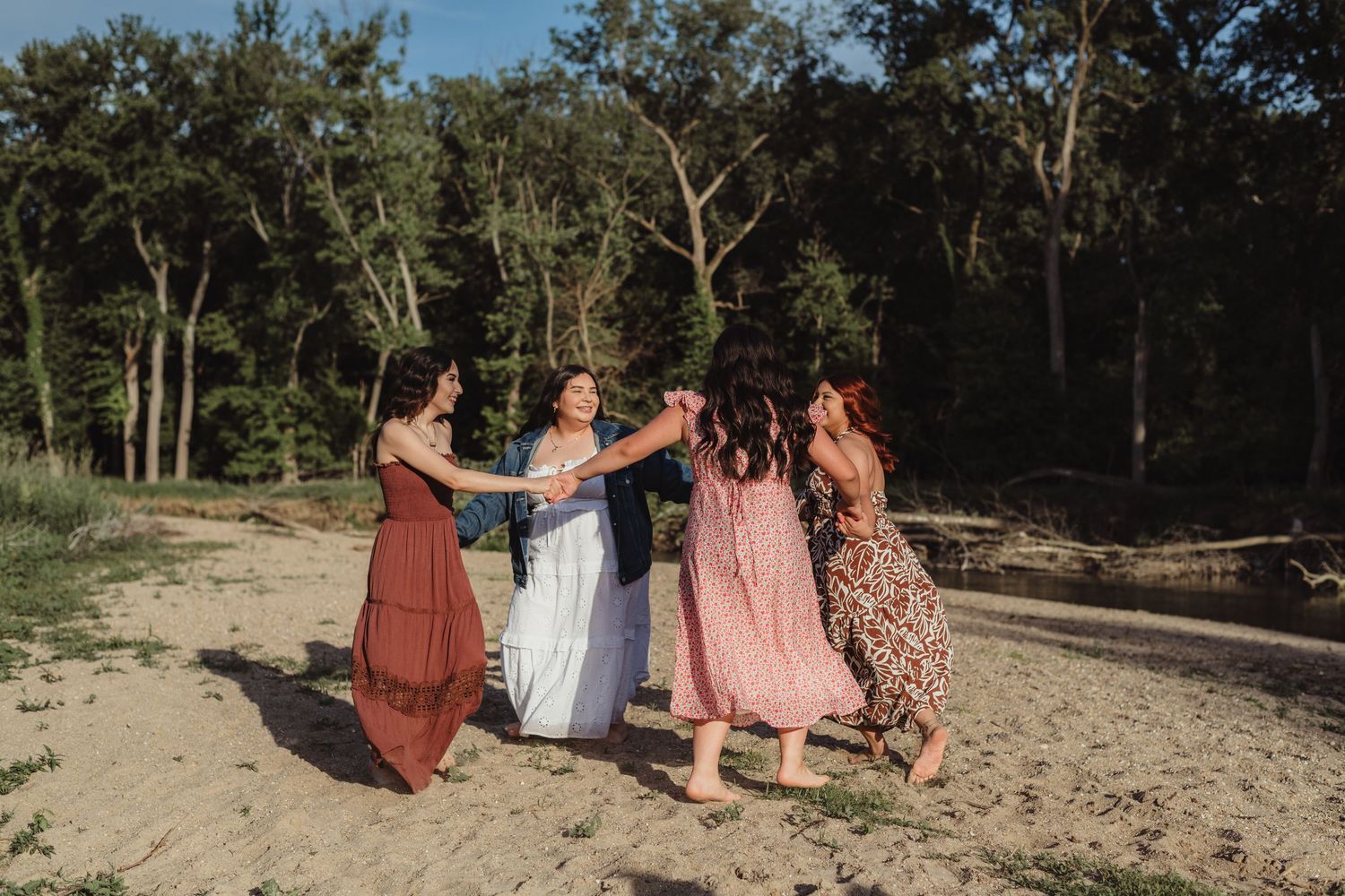 Five young women in flowy dresses dancing together on a sandy beach surrounded by trees.