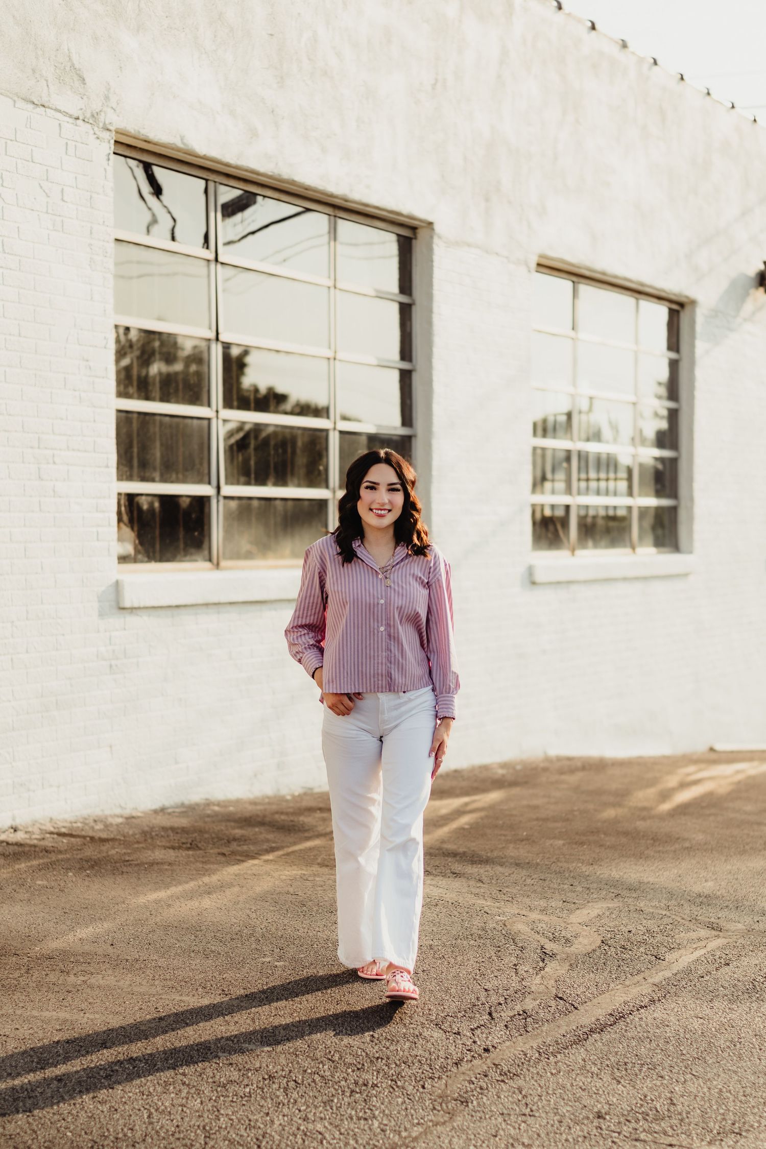 Woman in pink blouse and white flared jeans smiling outdoors near white brick building with large windows.