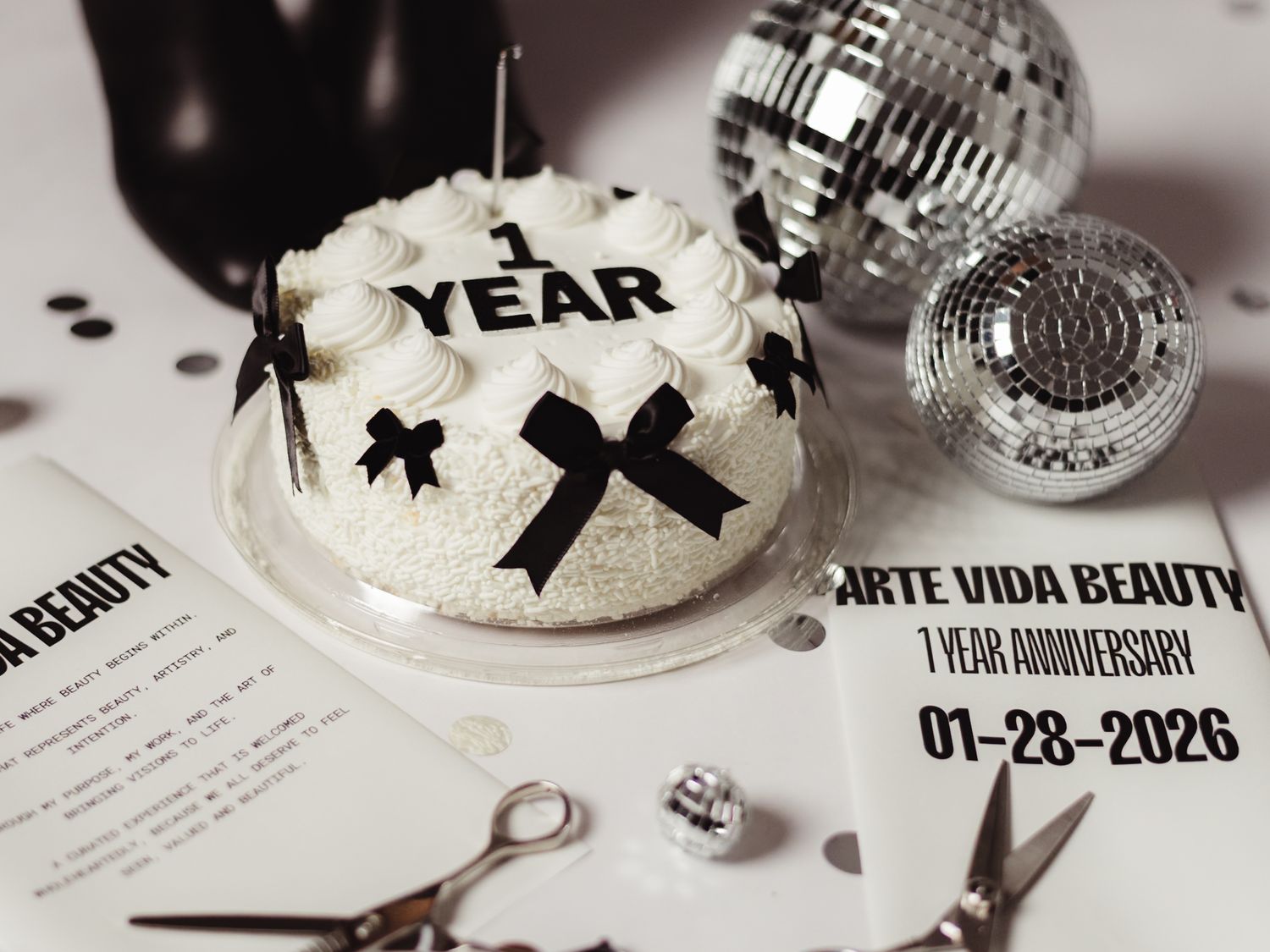 White anniversary cake with black bows and '1 Year' text, disco balls, and Arte Vida Beauty 1-year anniversary signage.