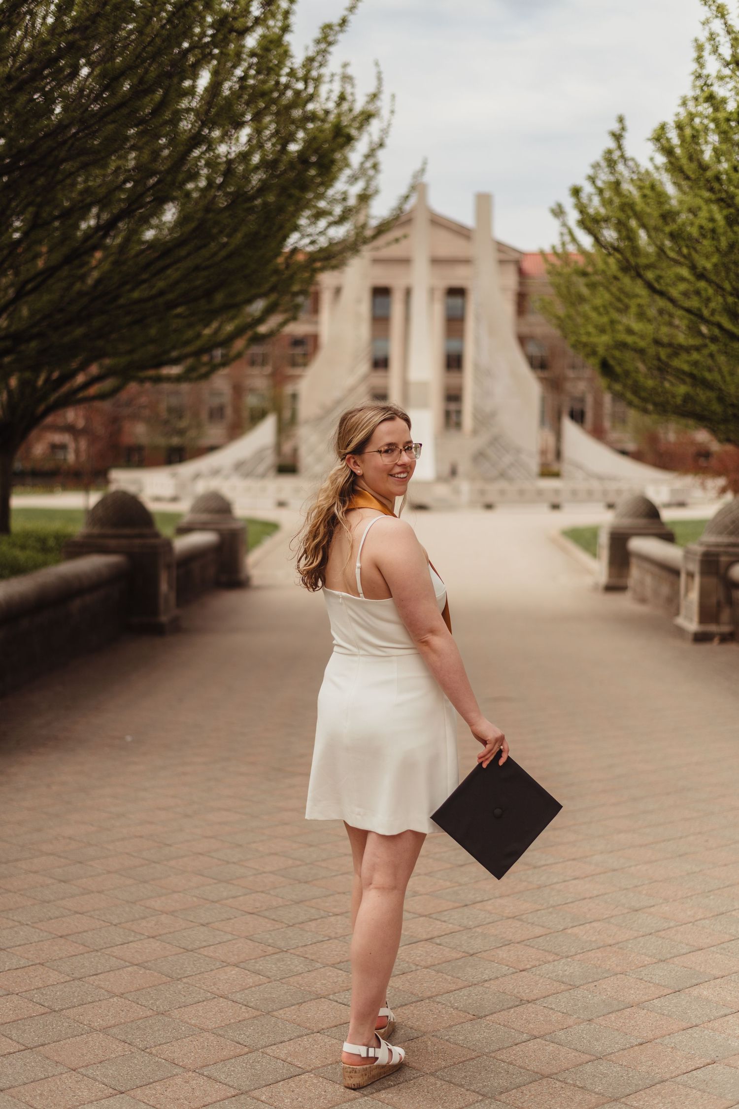 Graduate in white dress holds cap while walking on  on Purdue Campus in West Lafayette, IN