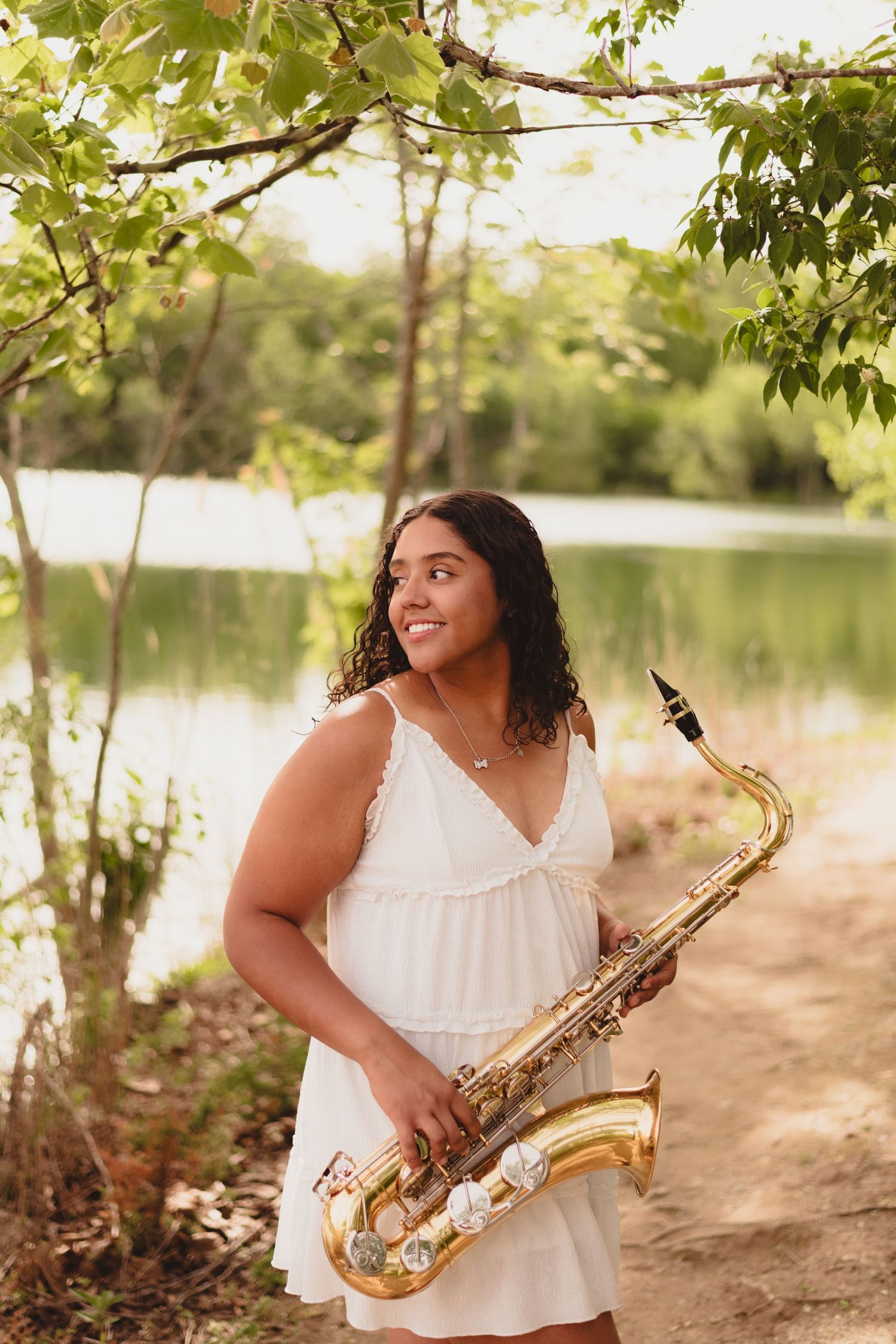 Musician in a white top plays saxophone outdoors near a serene lake during golden hour.