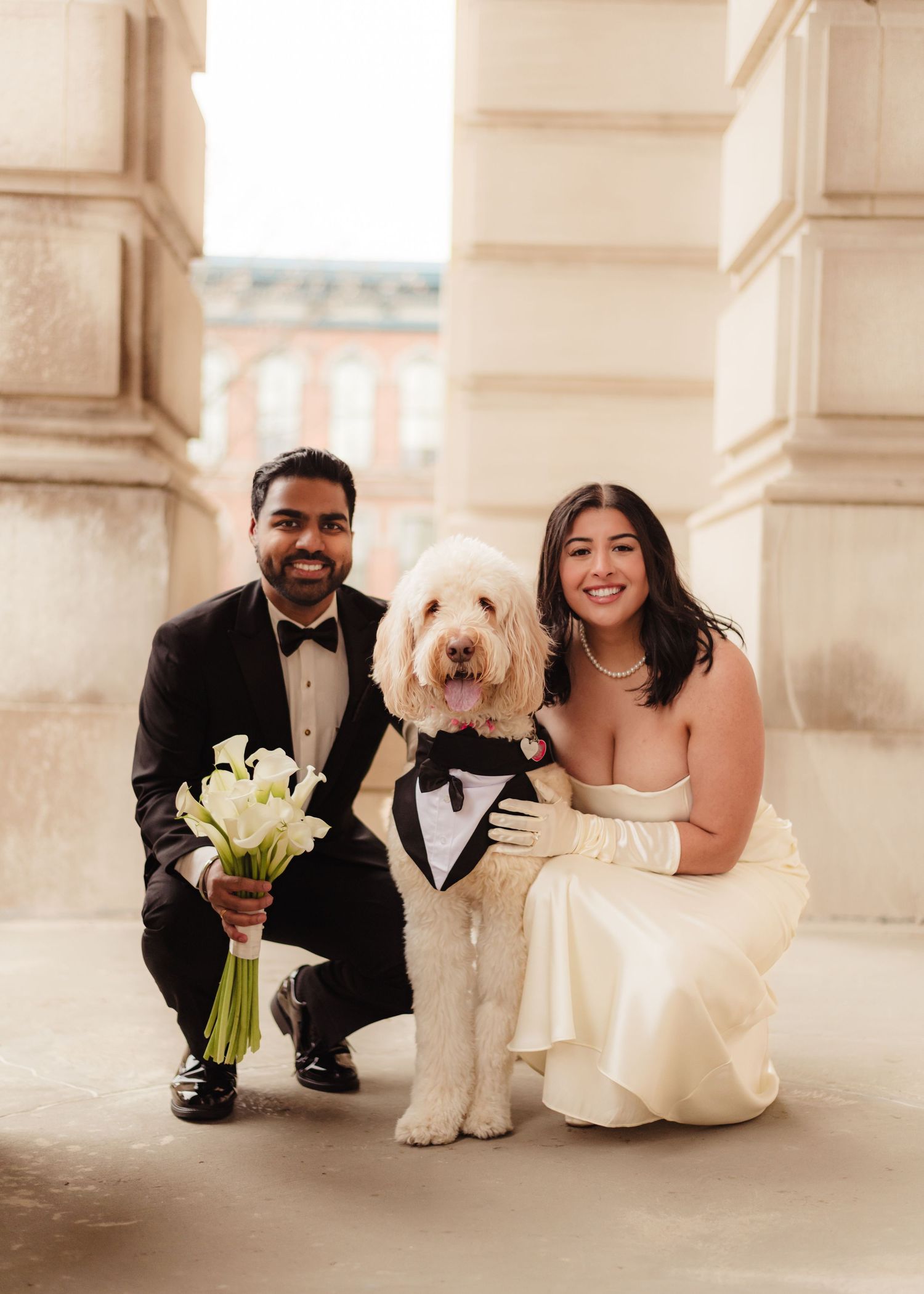 Wedding couple in formal attire poses with their white fluffy dog under elegant columns.