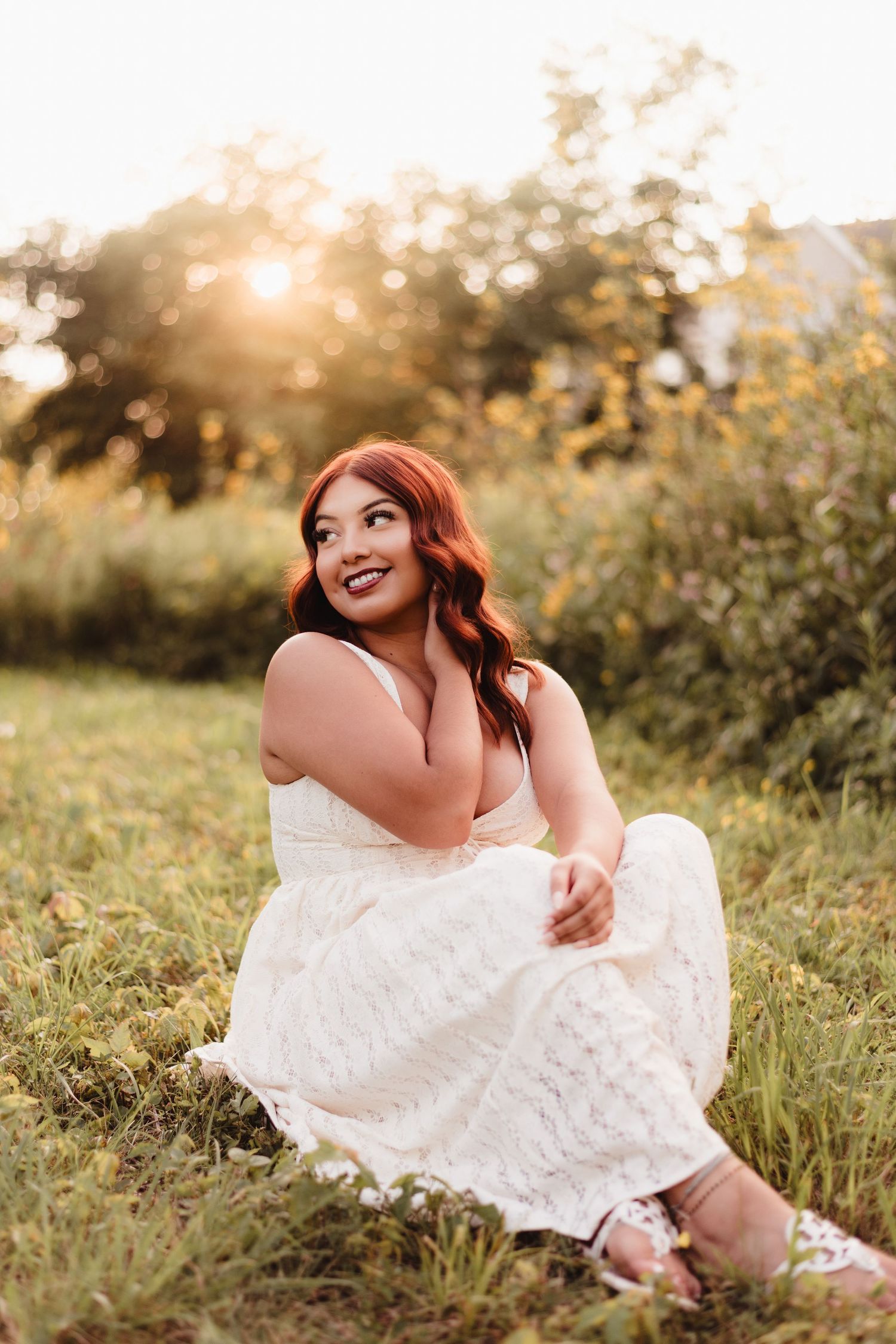 A smiling woman with red hair sits in a sunlit field wearing a white dress, glowing in golden hour light.