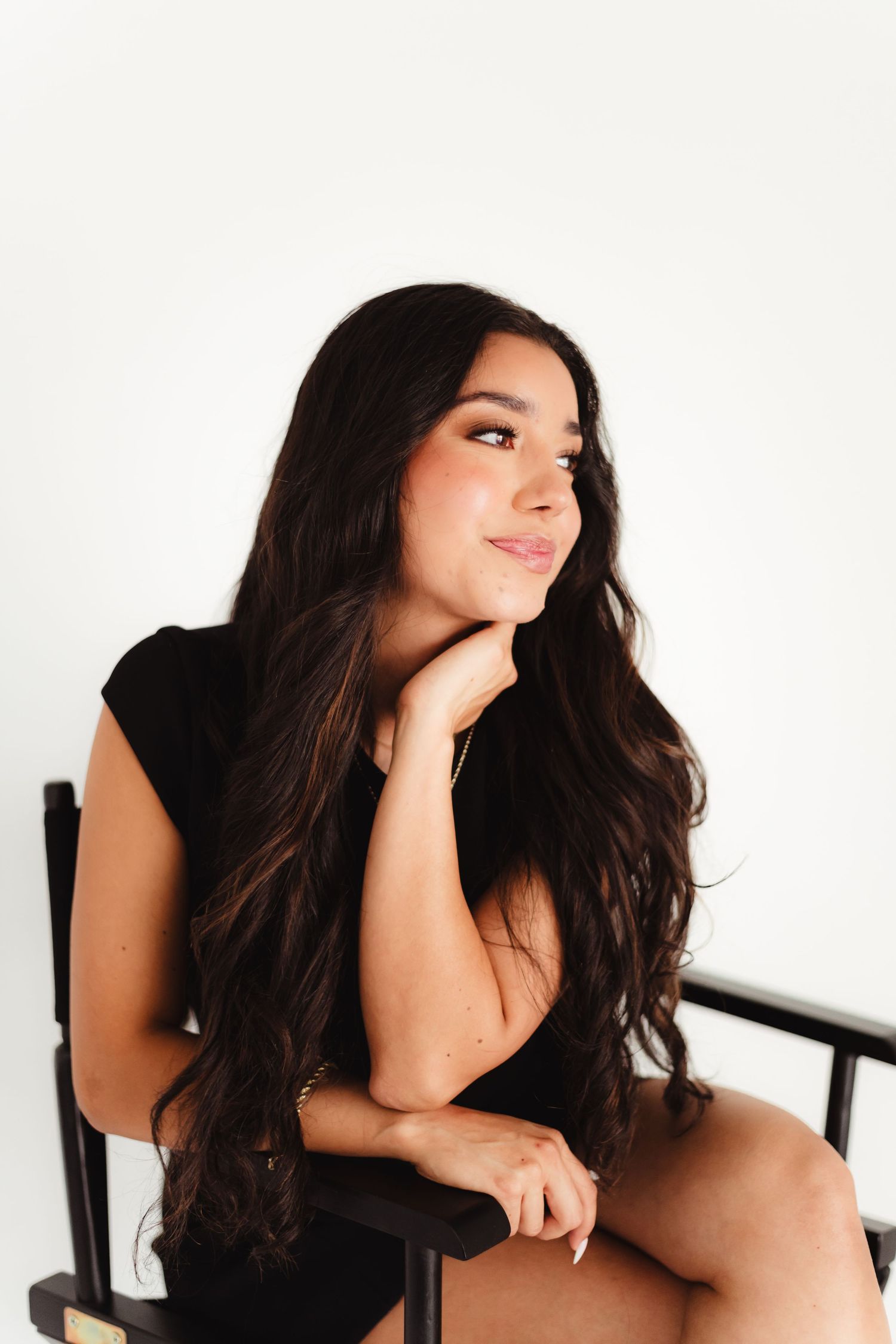 Young woman with long dark wavy hair sits in a director's chair, smiling thoughtfully against a white background.