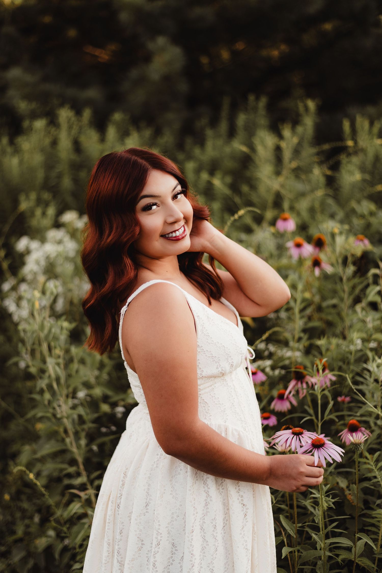 Smiling young woman with red hair in white lace dress holding pink flowers in a lush garden setting.