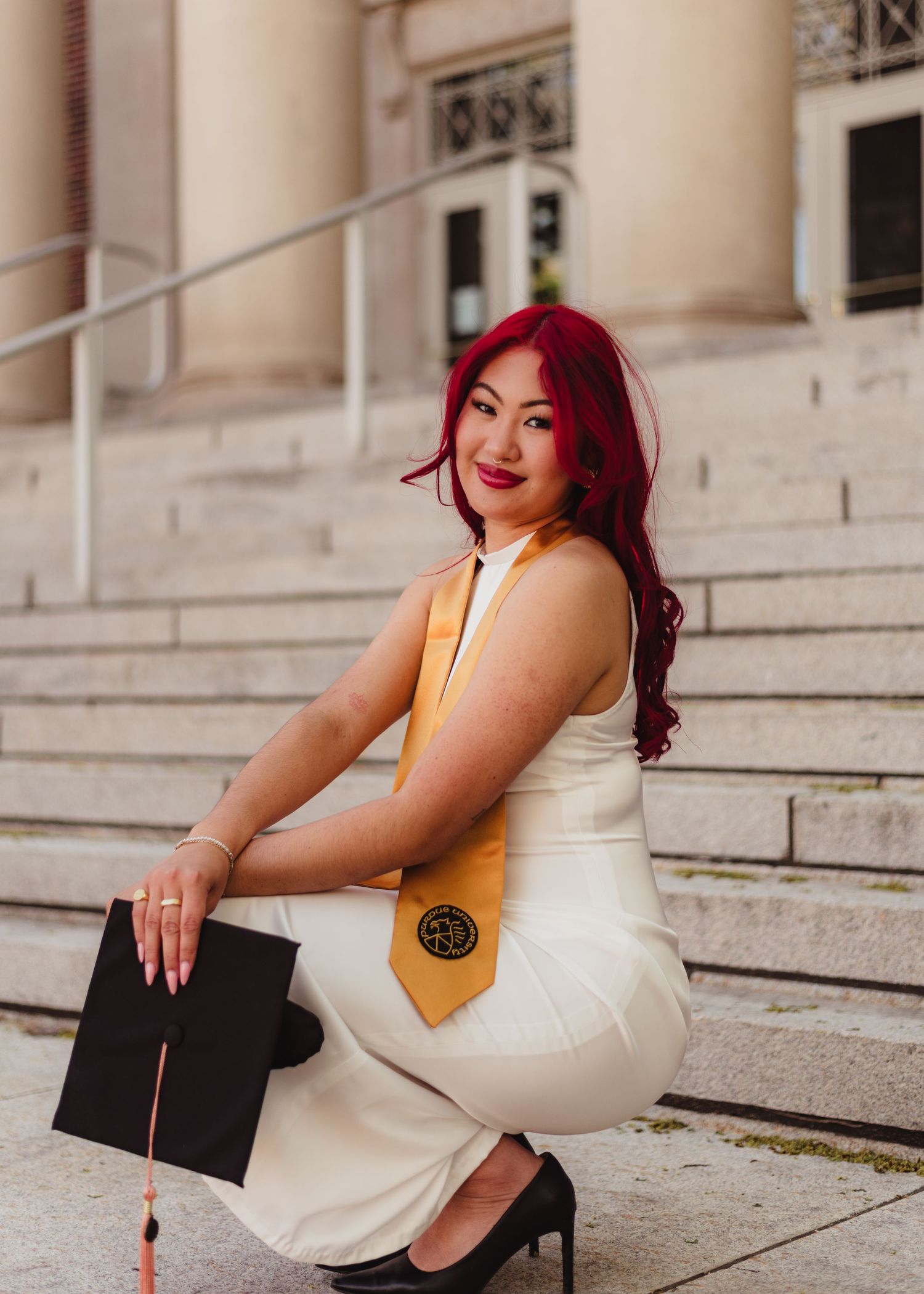 Graduate in white dress with red hair holds diploma while sitting on architectural stone steps at Purdue Campus in West Lafayette, IN