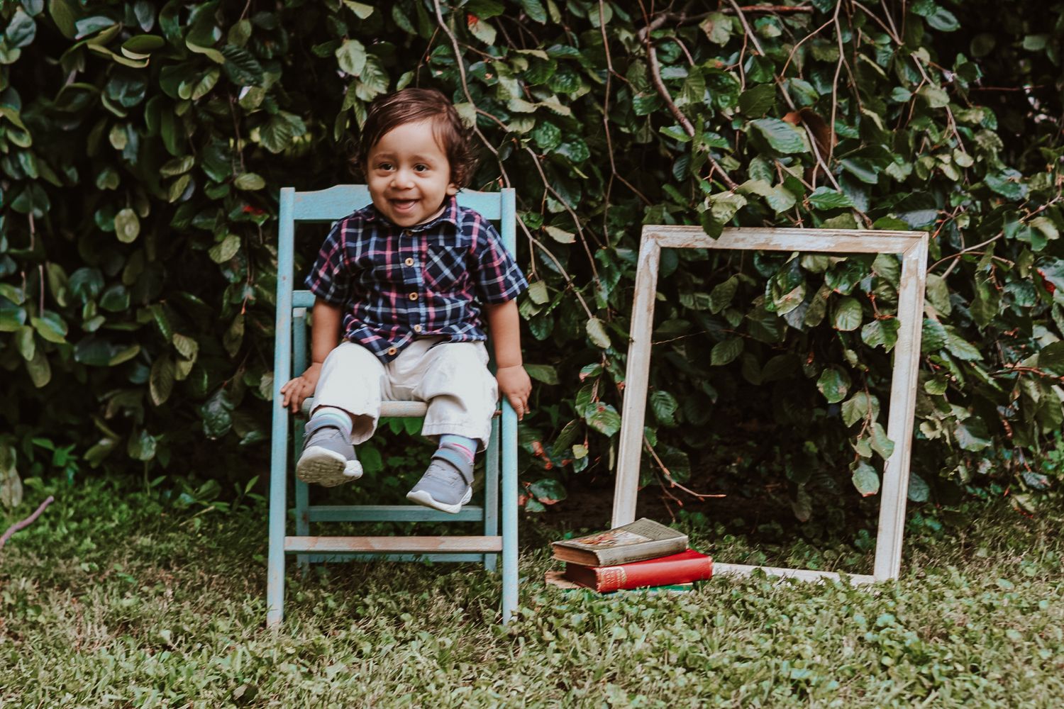 Wide shot of baby in plaid outfit sitting on blue chair against garden backdrop.