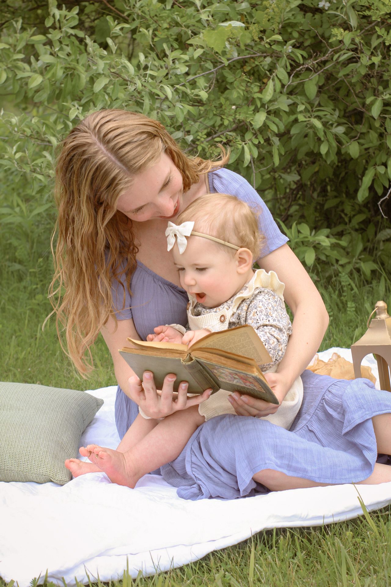 Woman in a blue dress enjoys a picnic on a white blanket with lanterns and old vintage books with her daughter. Cottage core vibes