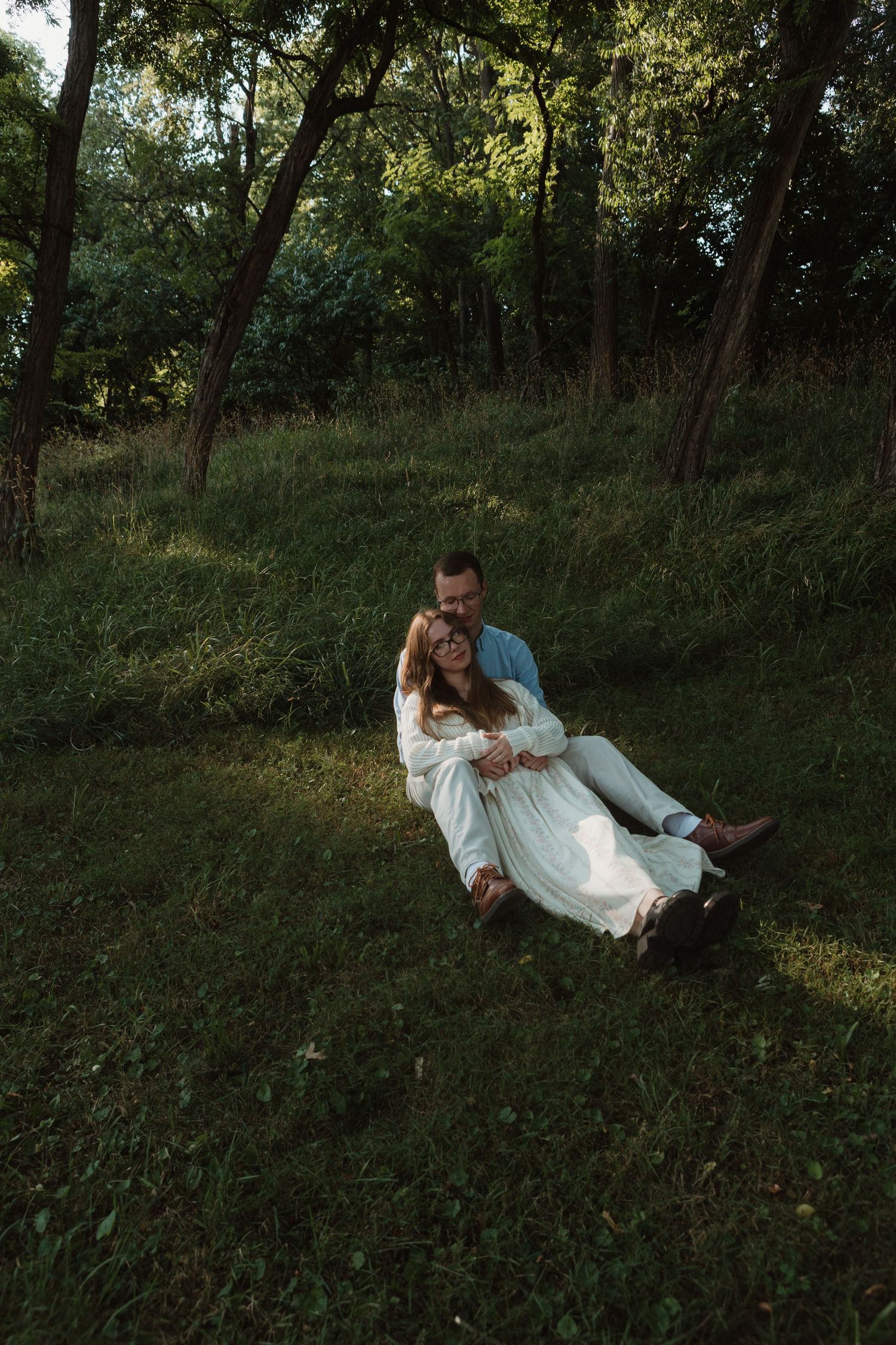 Couple sitting together on a grassy hillside surrounded by lush green trees in a romantic outdoor setting.