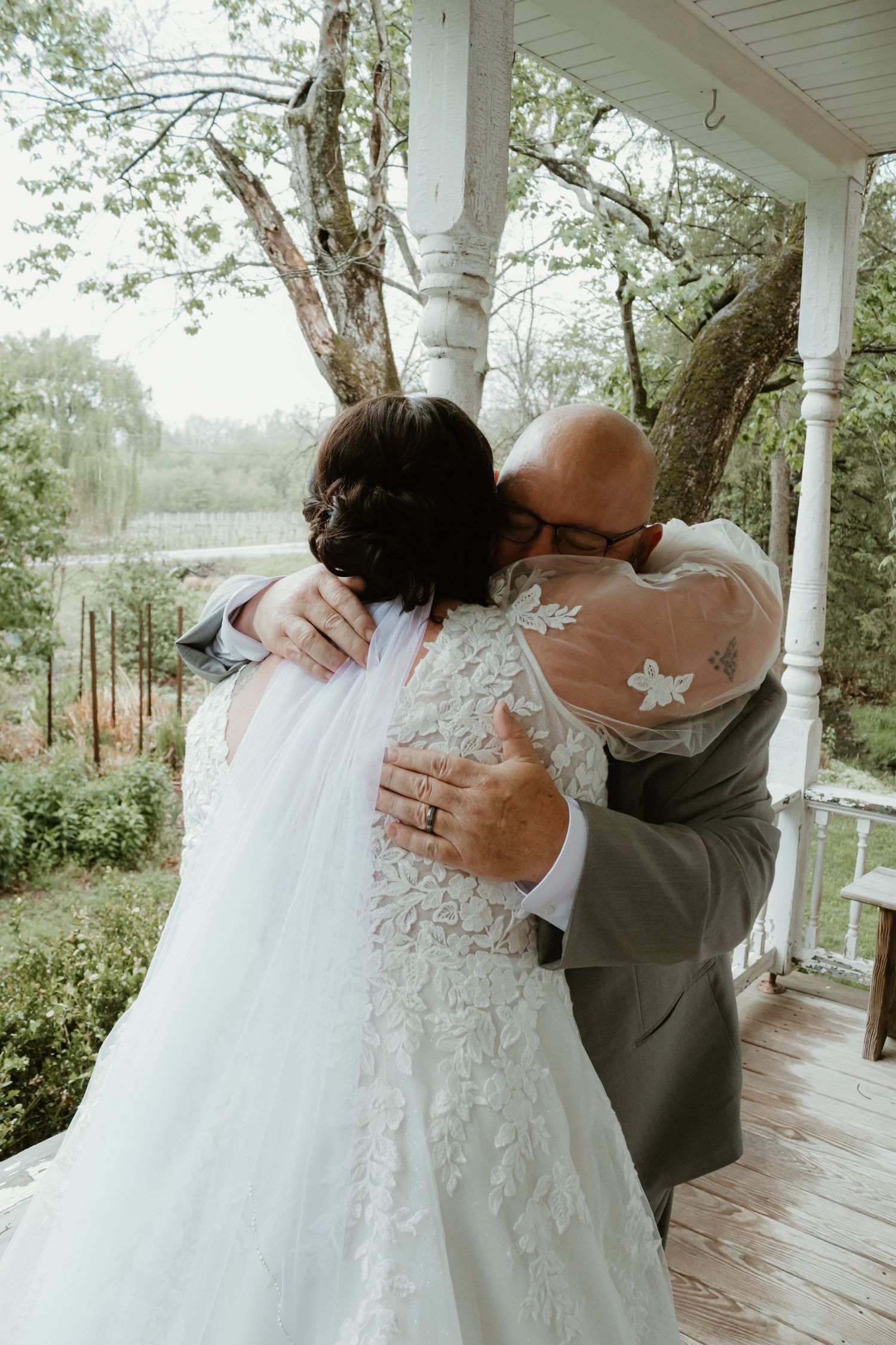 Bride in lace wedding gown embracing a man in a gray suit on a porch surrounded by lush greenery.