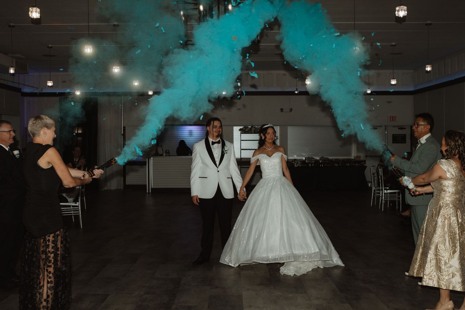 Bride and groom enter wedding reception with teal smoke cannons creating dramatic arch in elegant venue.