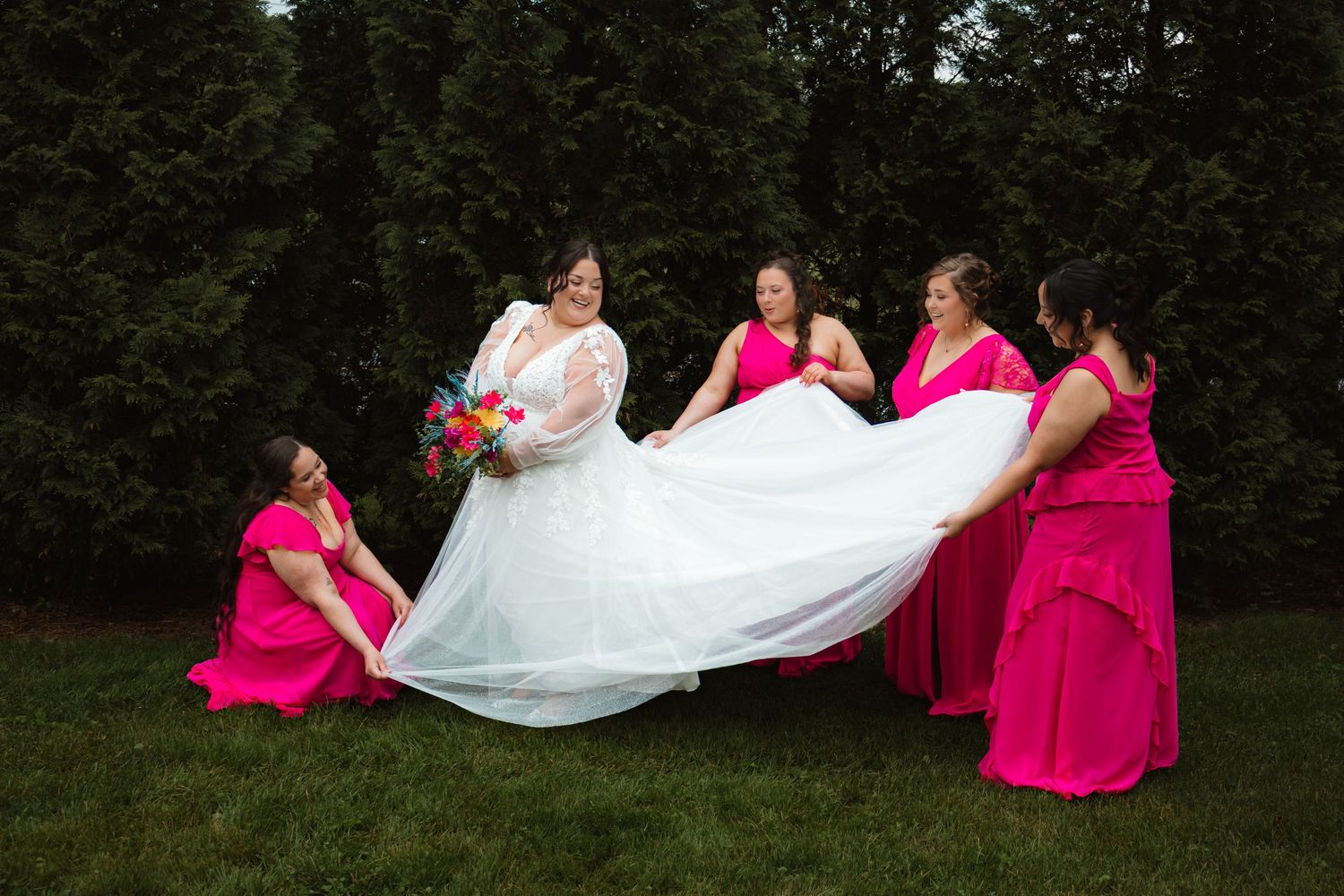 Bride in white gown with bridesmaids in hot pink dresses fluffing her skirt on green lawn.