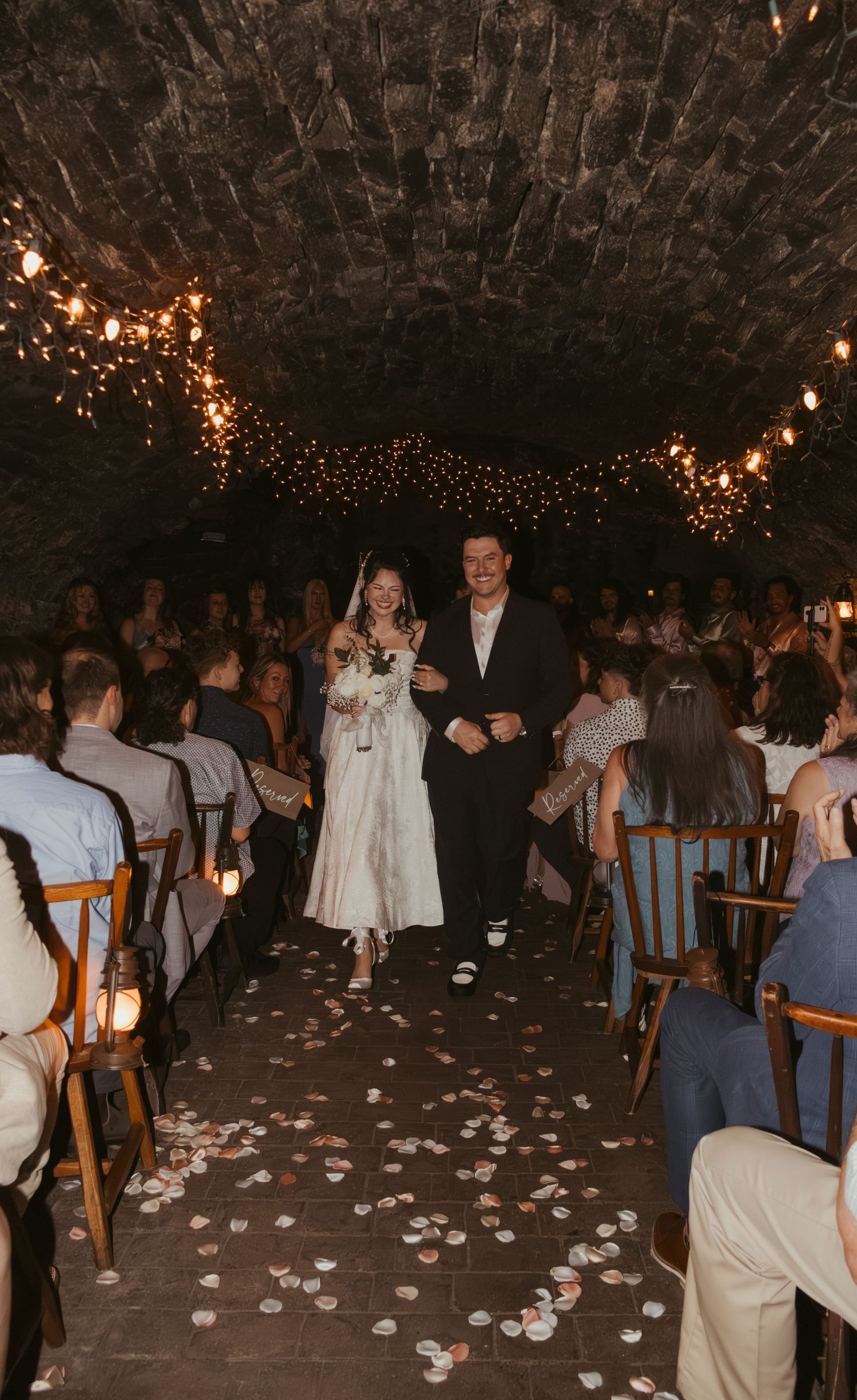 Bride and groom walk down flower petal aisle in romantic stone venue with string lights and seated wedding guests.