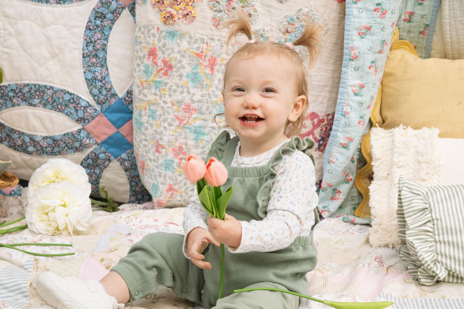 Smiling baby girl in green overalls holding pink tulips, sitting among colorful quilts and pillows.