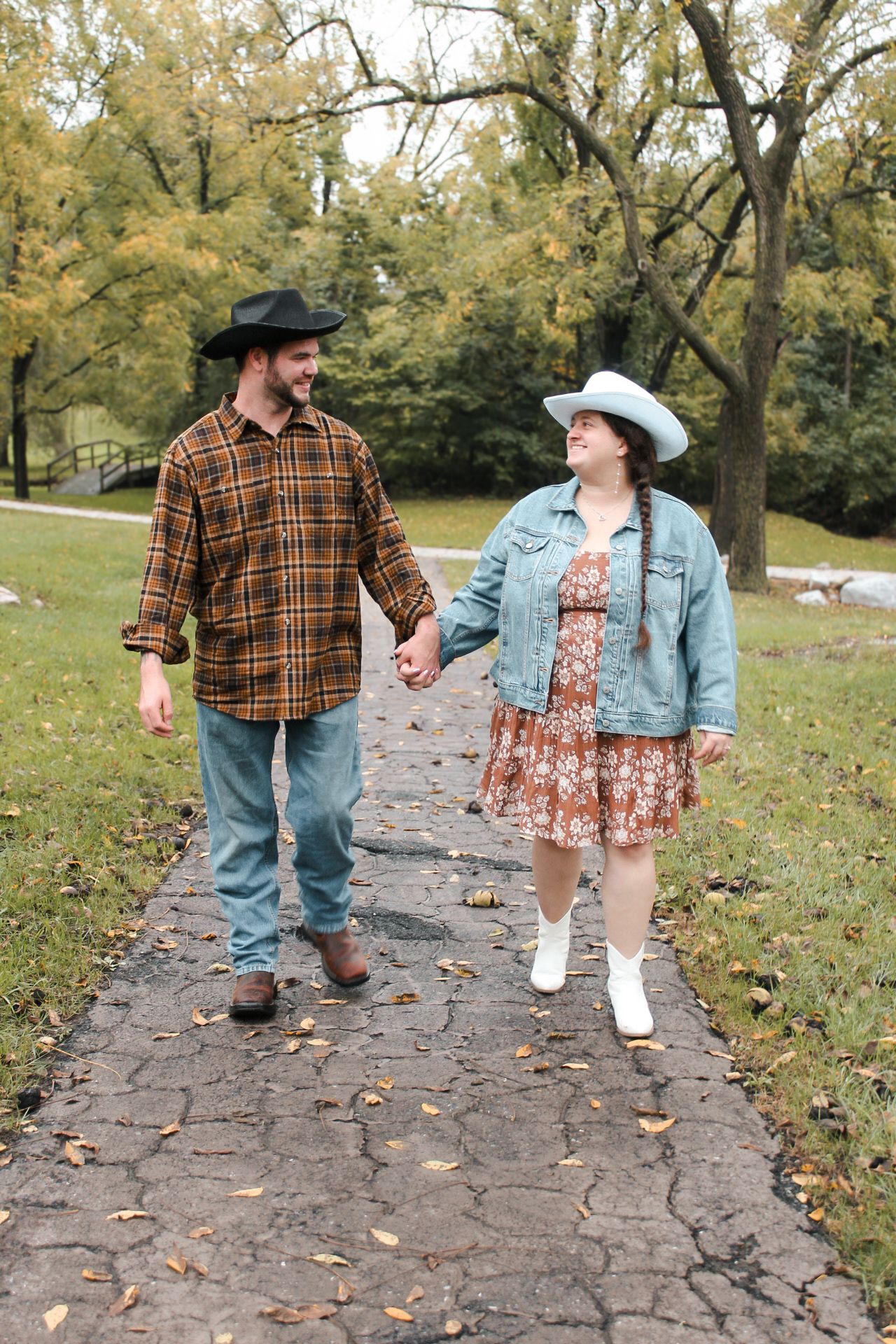 A couple walks hand in hand along a stone path in autumn, wearing western-style hats and casual attire.