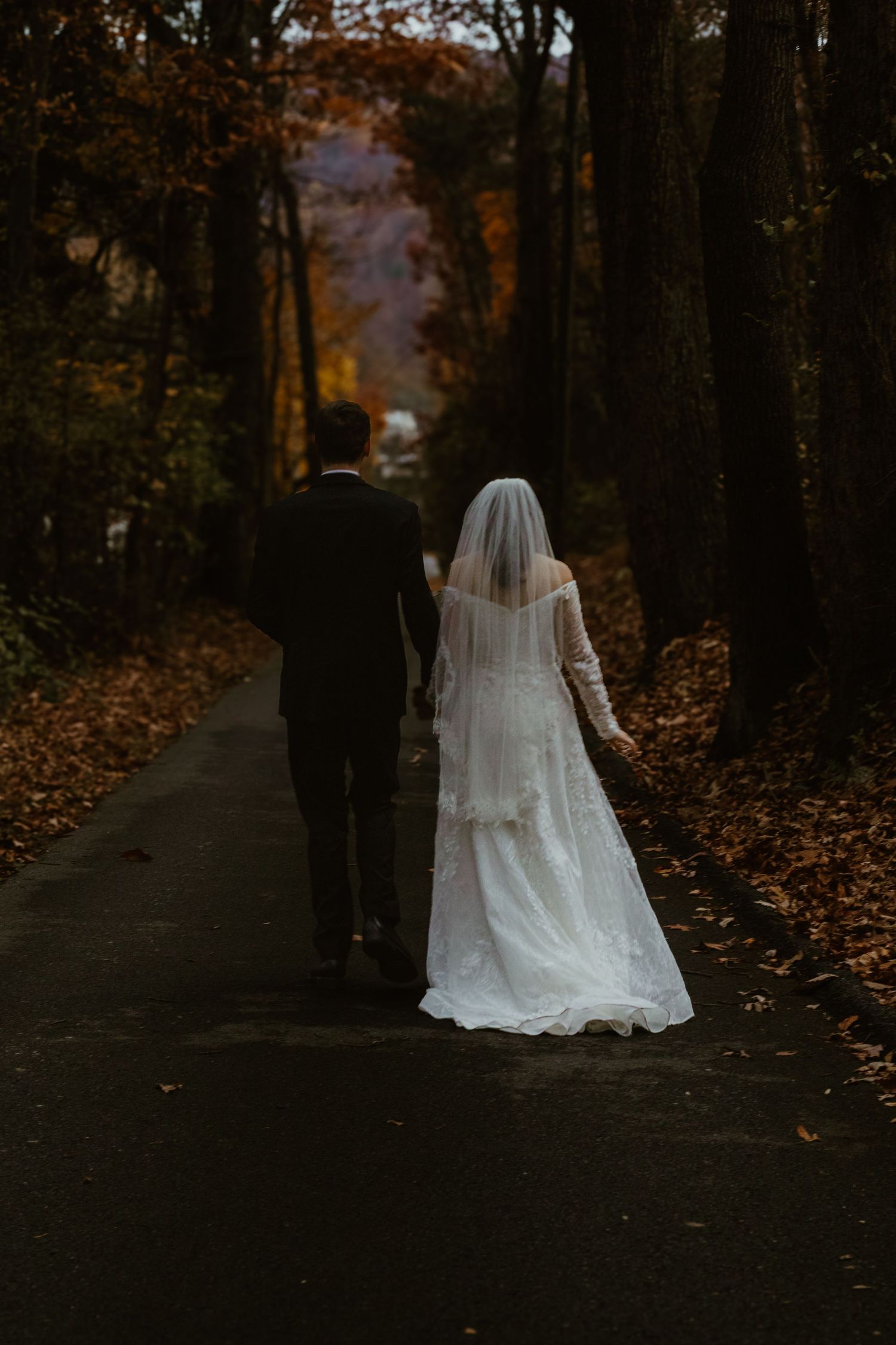 A wedding couple walks through a wooded path covered in fallen autumn leaves at dusk.
