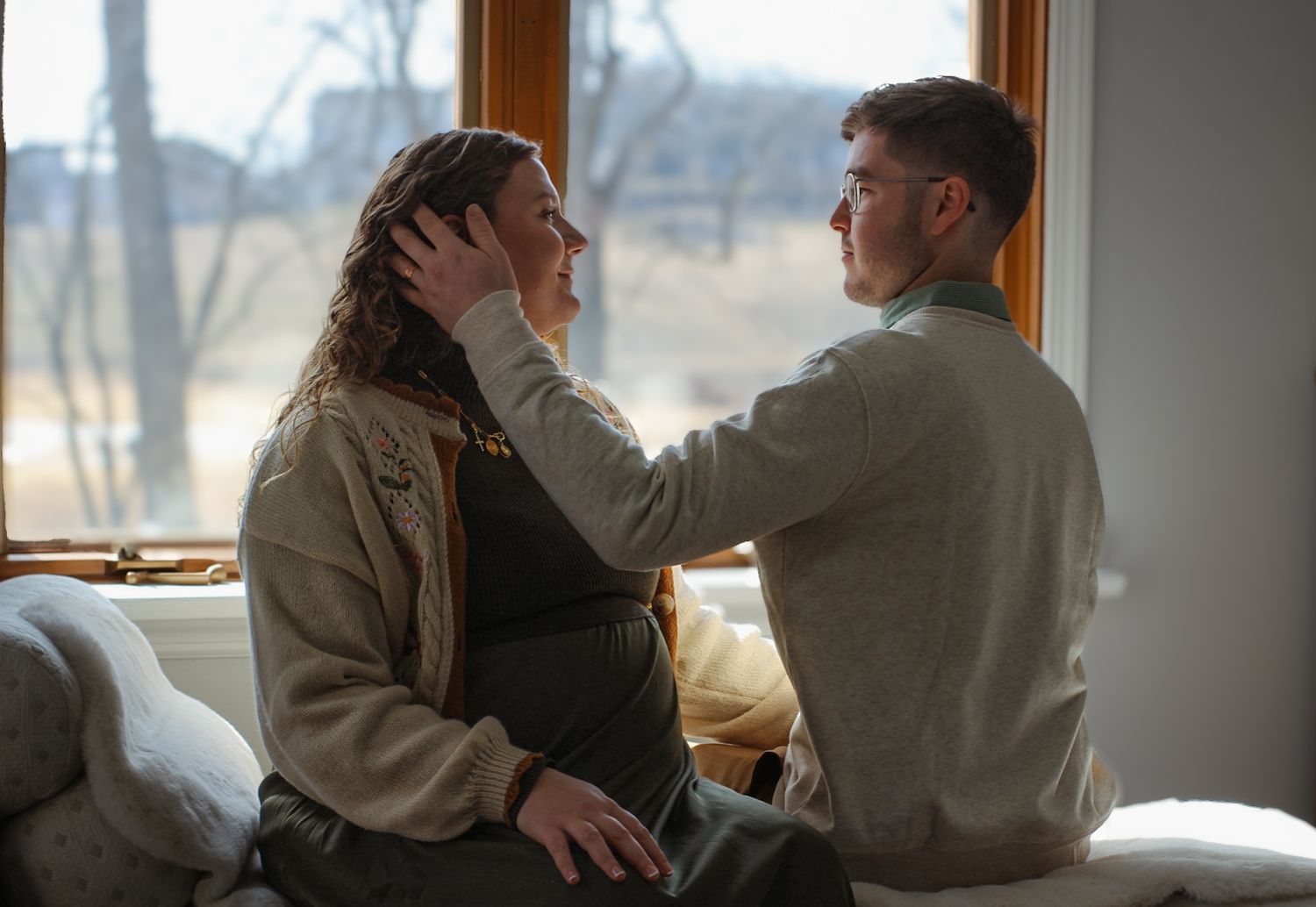 Two people in cozy sweaters share a tender moment by a window.