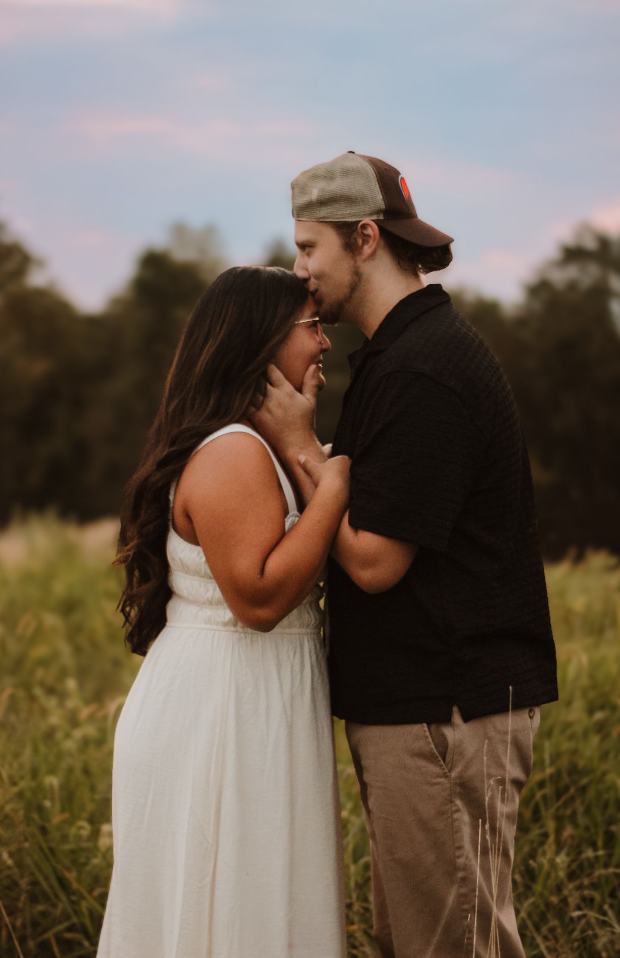A couple shares an intimate moment together in a field at sunset while wearing casual and white summer attire.