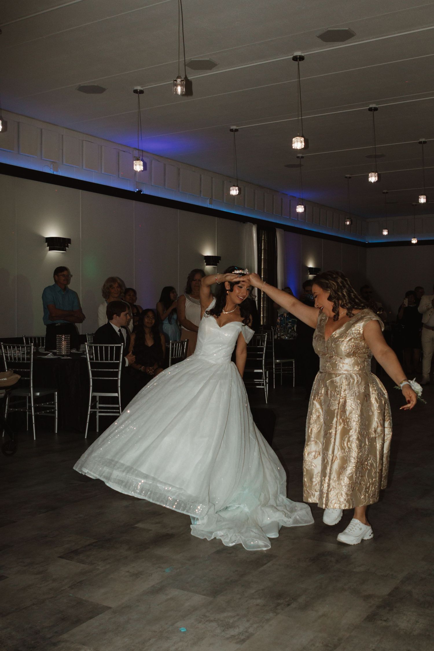 Bride in white gown dancing with woman in gold dress at elegant wedding reception venue.