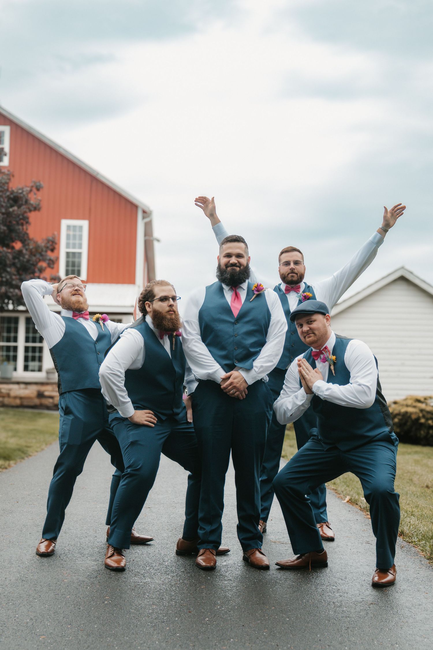 Five groomsmen in navy vests and colorful ties pose playfully outside a red barn on a wedding day.
